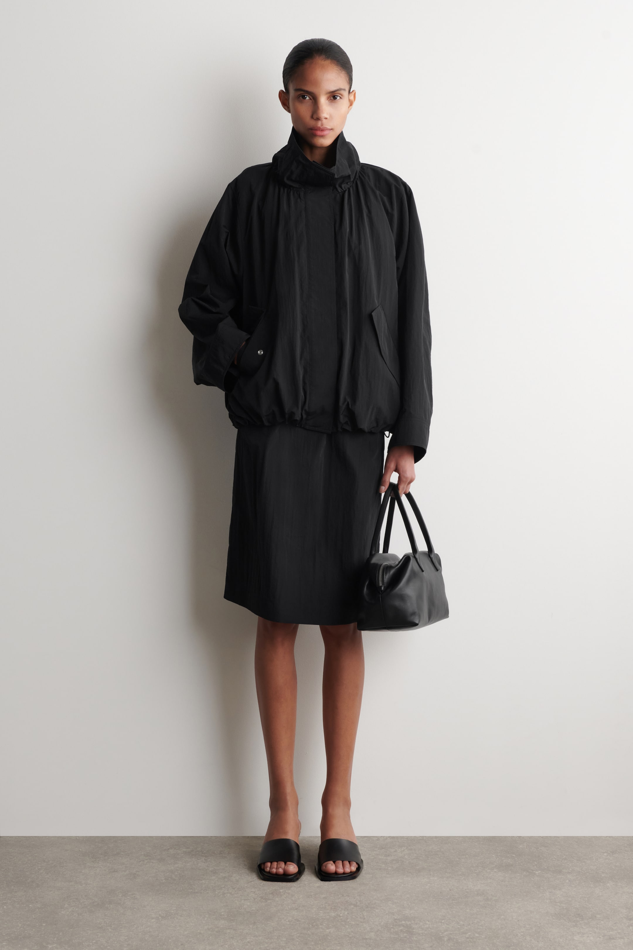 Agrandir l'image: Une veste noire texturée, dotée d'un col montant plissé, de manches longues décontractées, de poches latérales et d'un ourlet élastiqué, est portée avec une jupe midi noire. Un sac à main en cuir noir est porté, et des sandales à enfiler à bout carré noir sont portées.