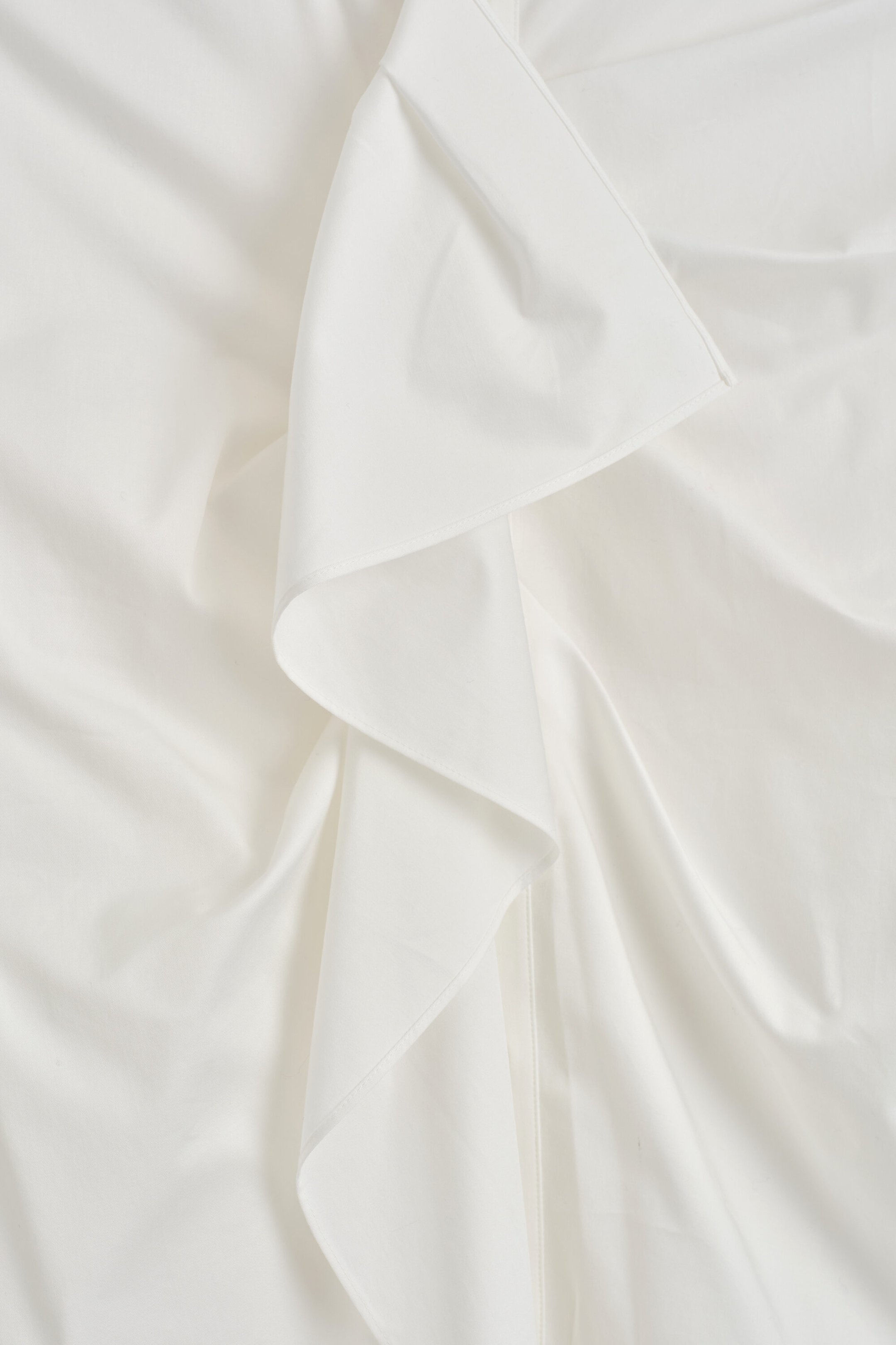 A close-up view of a white cotton blouse showing the soft, cascading ruffle detail along the placket. The fluid, lightweight fabric drapes in soft folds to highlight the clean-finished edges and delicate structure of the ruffle.