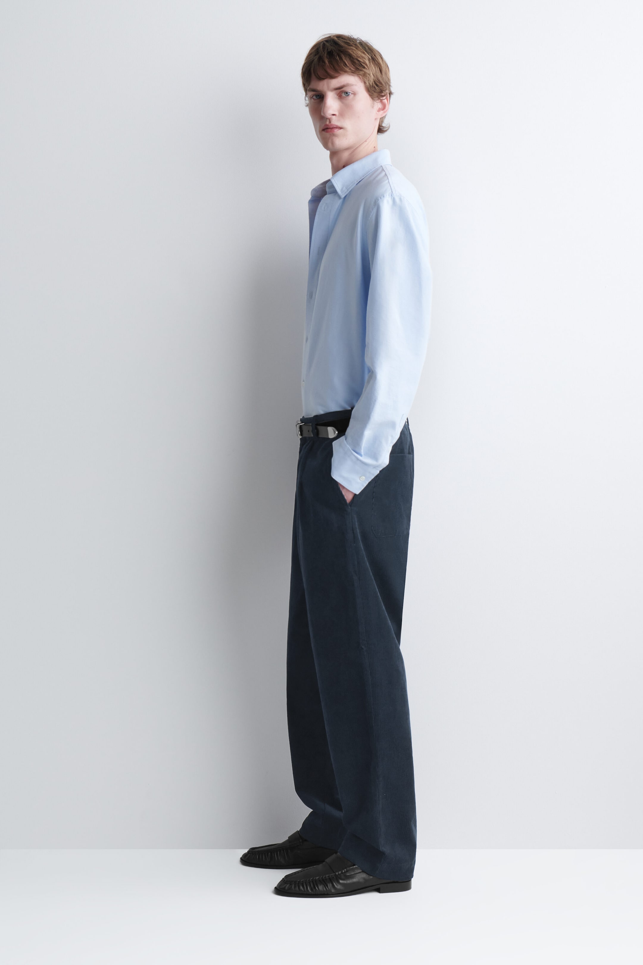 Corduroy relaxed-fit tapered pants in dark navy blue are worn by the model, paired with a tucked-in light blue long-sleeve shirt and a black leather belt. The pants feature a textured ribbed finish, side pockets, and a loose, tapered silhouette, with the model standing in profile and resting one hand in a pocket. Corduroy relaxed-fit tapered pants in dark navy blue are worn by the model, paired with a tucked-in light blue long-sleeve shirt and a black leather belt. The pants feature a textured ribbed finish, side pockets, and a loose, tapered silhouette, with the model standing in profile and resting one hand in a pocket.