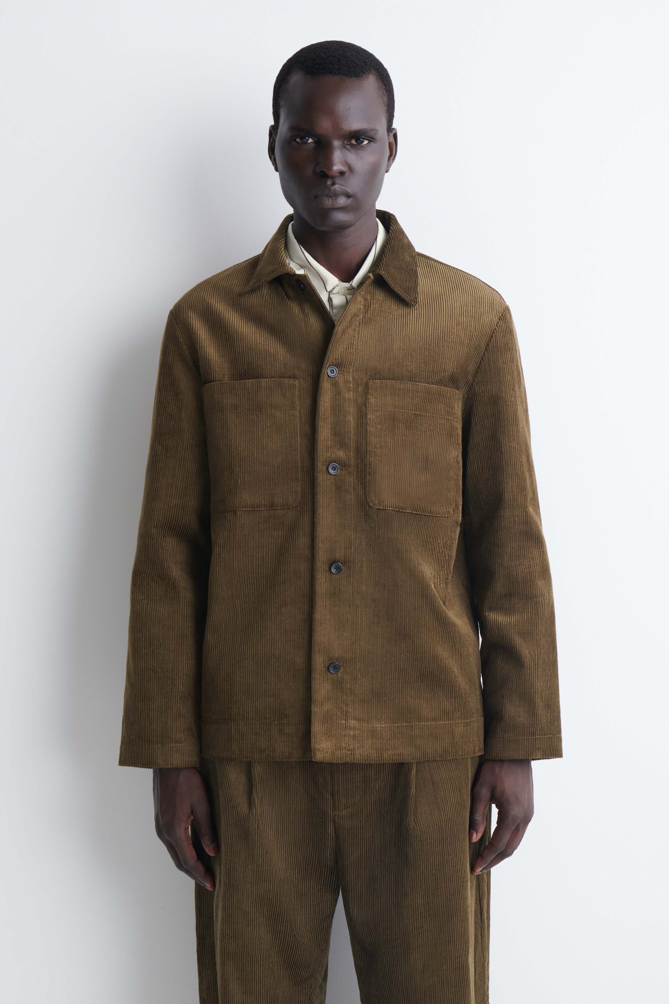 View larger image: This front view presents a brown cotton corduroy overshirt featuring a pointed collar, button-front fastening, and two large chest patch pockets. The overshirt is layered over a light-coloured collared shirt and paired with matching corduroy trousers, highlighting its relaxed fit and ribbed texture.