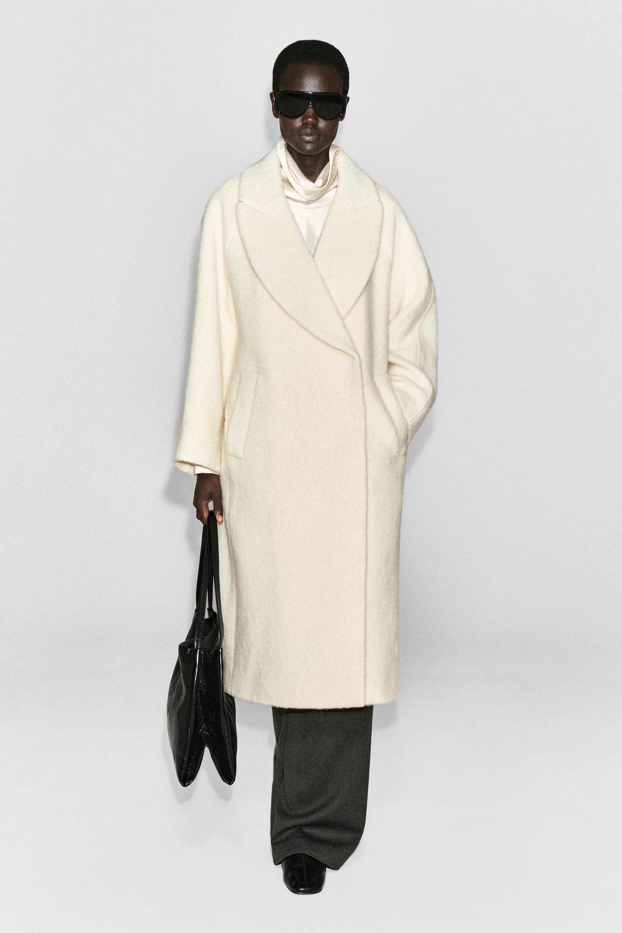 Agrandir l'image: Un manteau maxi en laine couleur crème, agréable au toucher, est porté de face, mettant en valeur sa coupe décontractée et sa finition texturée brossée. Le design présente de larges revers et des poches latérales en biais, superposé à un top à col montant et à un pantalon foncé.