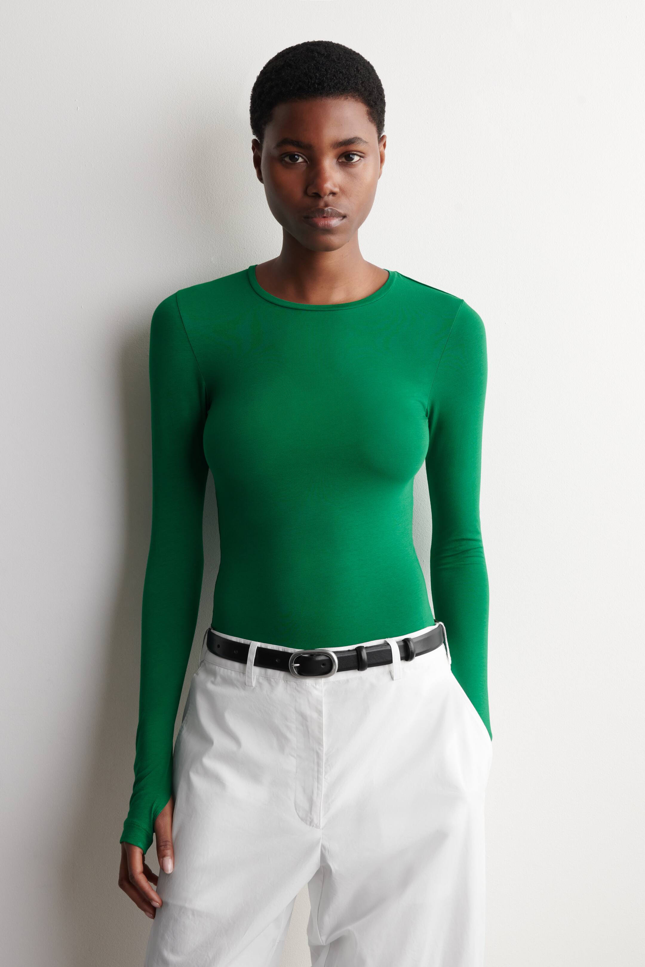 Grösseres Bild ansehen: Das grüne Langarmshirt wird in der Vorderansicht präsentiert, mit Rundhalsausschnitt, einer figurbetonten Silhouette und einem Daumenloch-Detail am linken Ärmel, während es in weiße Hosen gesteckt getragen wird.