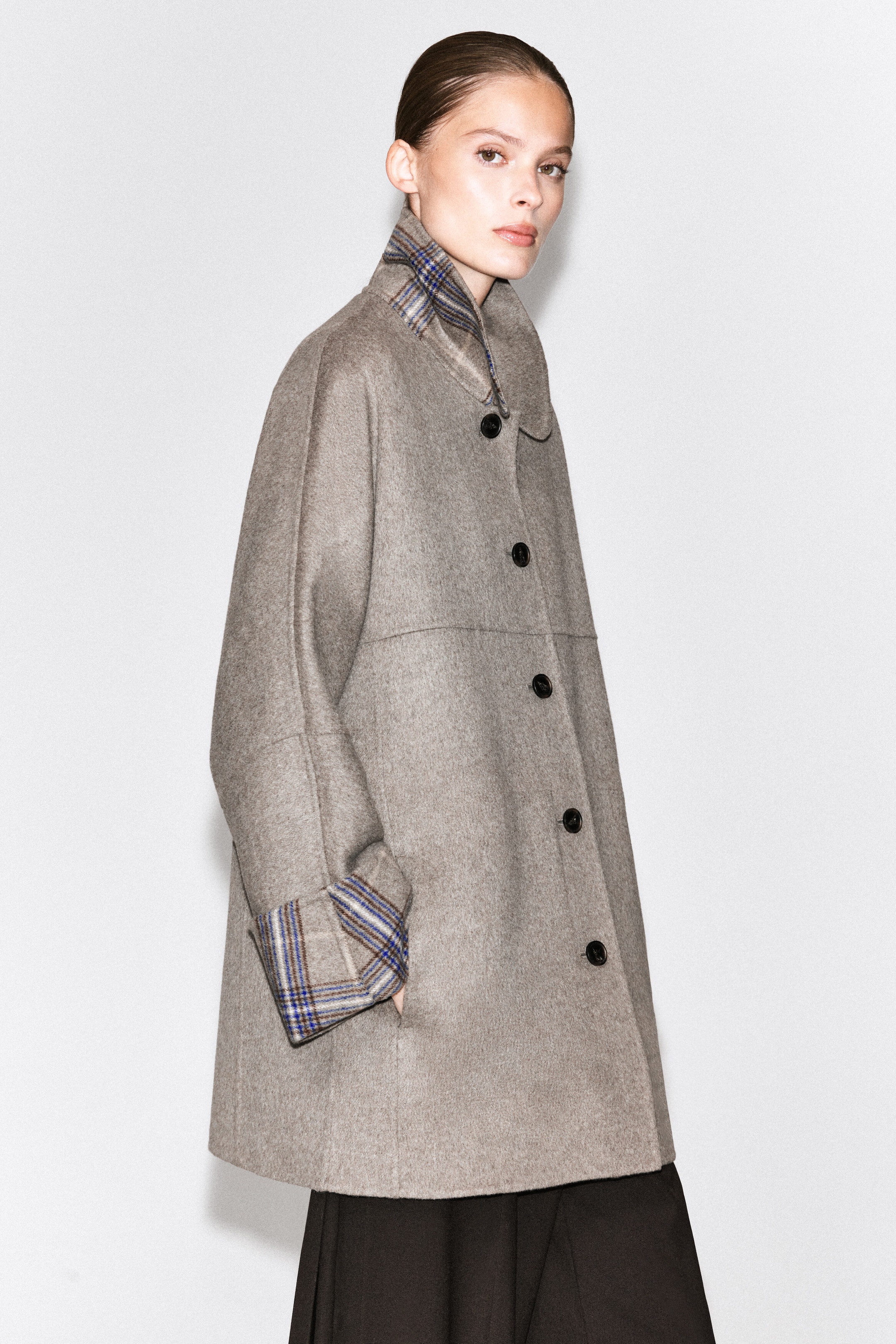 The model is wearing a beige, double-faced coat with a button-front closure, a raised collar, and folded cuffs that reveal a blue and brown check pattern. The model is standing with one hand in the coat's side pocket, styling it over dark trousers.