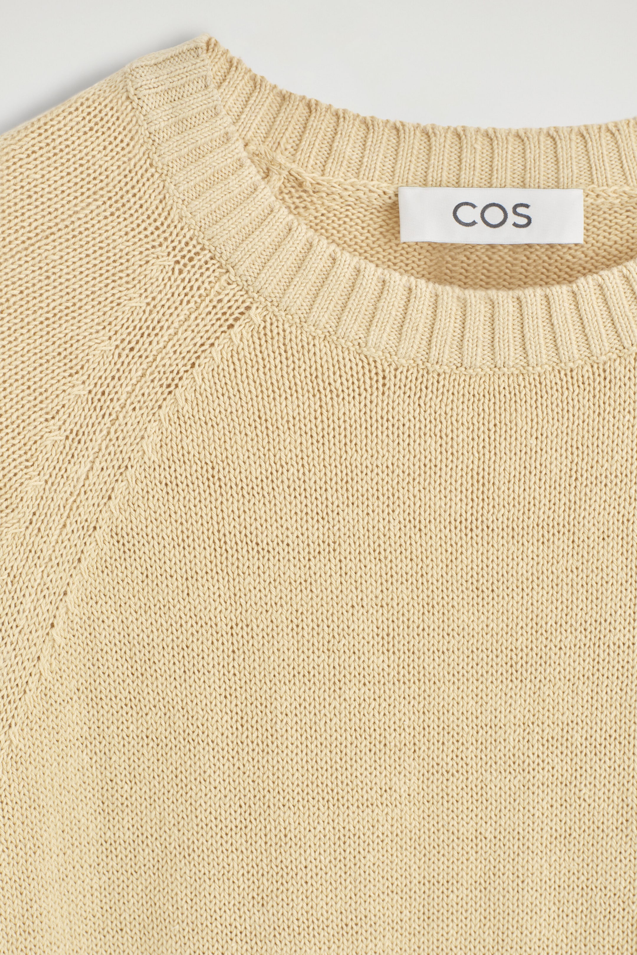 The Shrunken Knitted Linen T-shirt is shown in a close-up front view, highlighting its pale-beige knit texture and ribbed crew neck collar. It features raglan sleeve construction with openwork stitch detailing along the shoulder seam. The Shrunken Knitted Linen T-shirt is shown in a close-up front view, highlighting its pale-beige knit texture and ribbed crew neck collar. It features raglan sleeve construction with openwork stitch detailing along the shoulder seam.