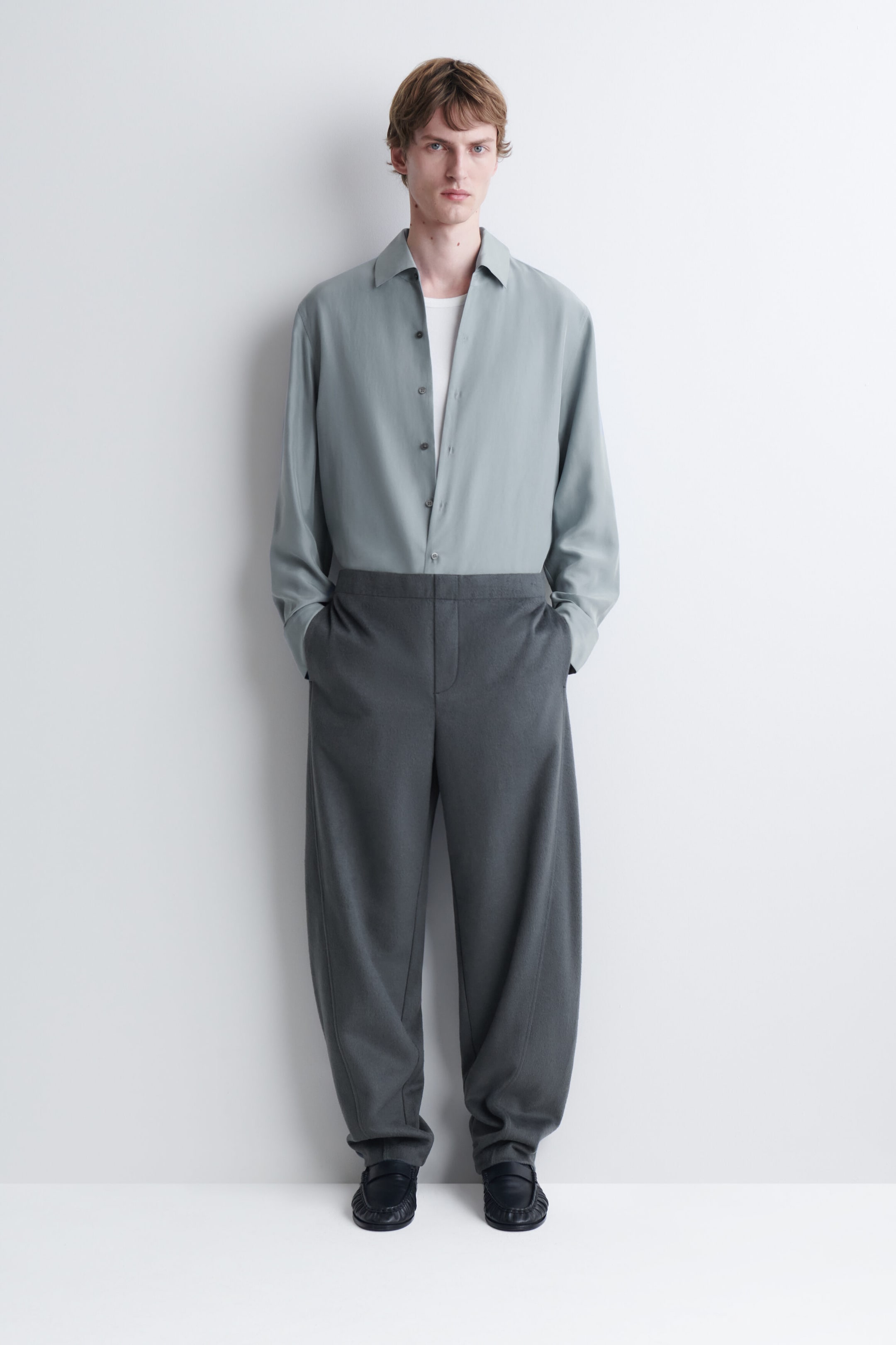 Le mannequin porte un pantalon gris à jambes larges et se tient debout, les mains dans les poches latérales. Le pantalon est agencé avec une chemise boutonnée à manches longues vert sauge pâle, rentrée à l’intérieur, superposée sur un t-shirt blanc.