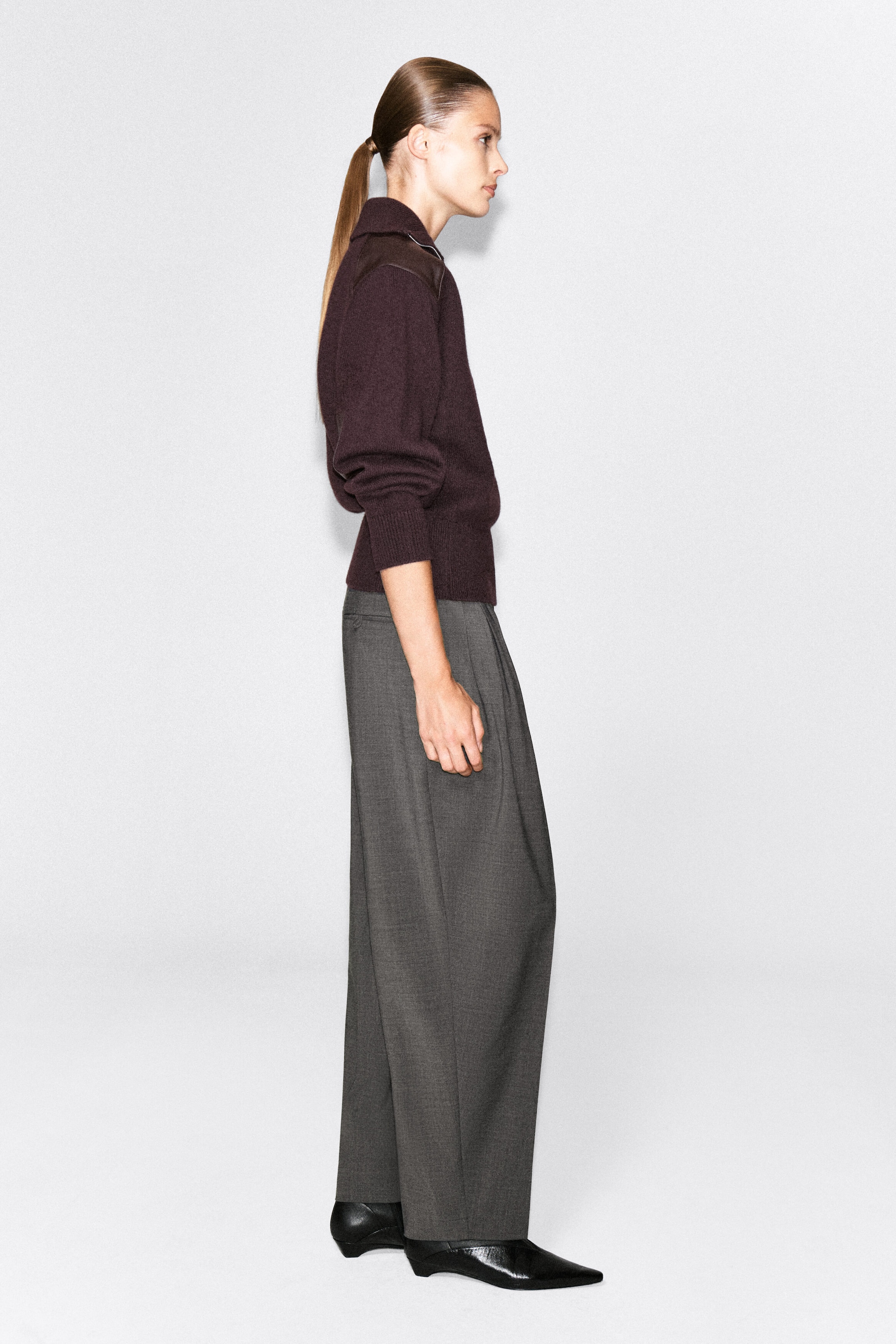 A dark plum merino wool zip-up sweater featuring a leather shoulder patch is being worn by the model, who is standing in profile. The sweater is styled with gray wide-leg trousers and black pointed-toe shoes. A dark plum merino wool zip-up sweater featuring a leather shoulder patch is being worn by the model, who is standing in profile. The sweater is styled with gray wide-leg trousers and black pointed-toe shoes.
