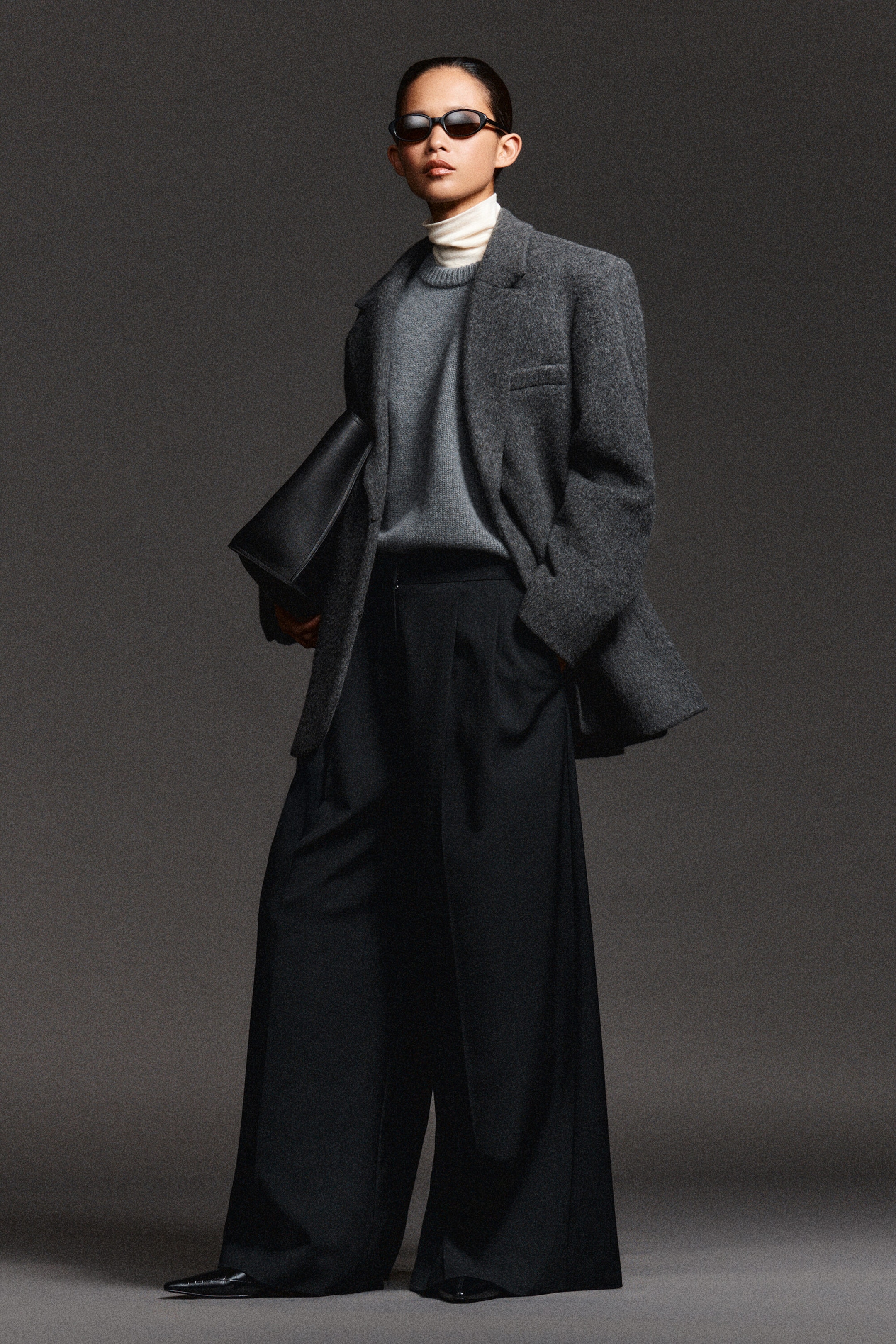 Pleated wide-leg trousers in black are shown from the front, featuring a high-rise waist with deep front pleats that create a draped, voluminous silhouette. The full-length legs pool slightly over pointed-toe shoes, with a grey knit jumper partially tucked into the waistband to highlight the structured waist design. Pleated wide-leg trousers in black are shown from the front, featuring a high-rise waist with deep front pleats that create a draped, voluminous silhouette. The full-length legs pool slightly over pointed-toe shoes, with a grey knit jumper partially tucked into the waistband to highlight the structured waist design.