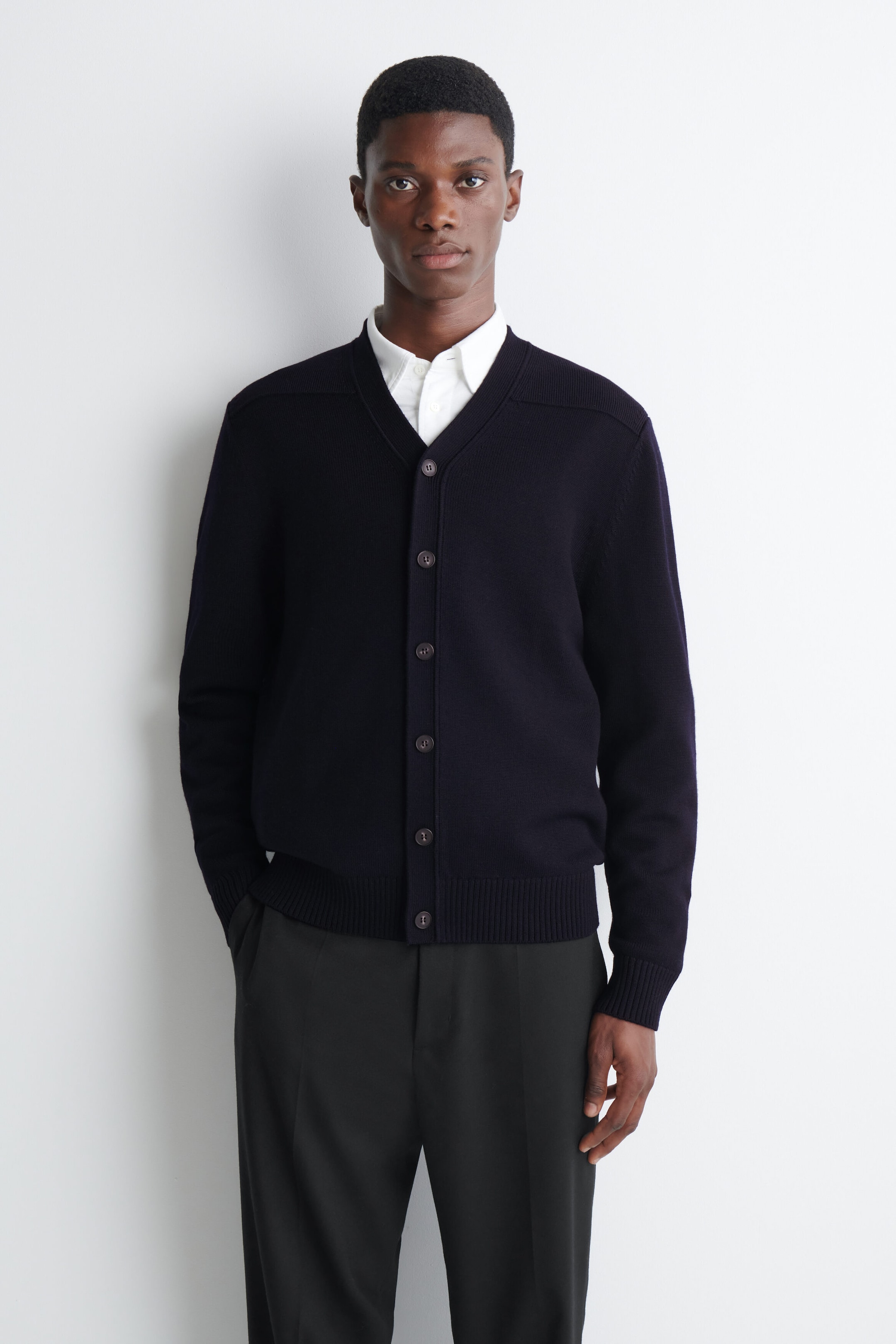A dark navy-blue V-neck cardigan with a front button fastening and ribbed trim is being worn by the model. The cardigan is layered over a white collared shirt and paired with dark tailored trousers. A dark navy-blue V-neck cardigan with a front button fastening and ribbed trim is being worn by the model. The cardigan is layered over a white collared shirt and paired with dark tailored trousers.
