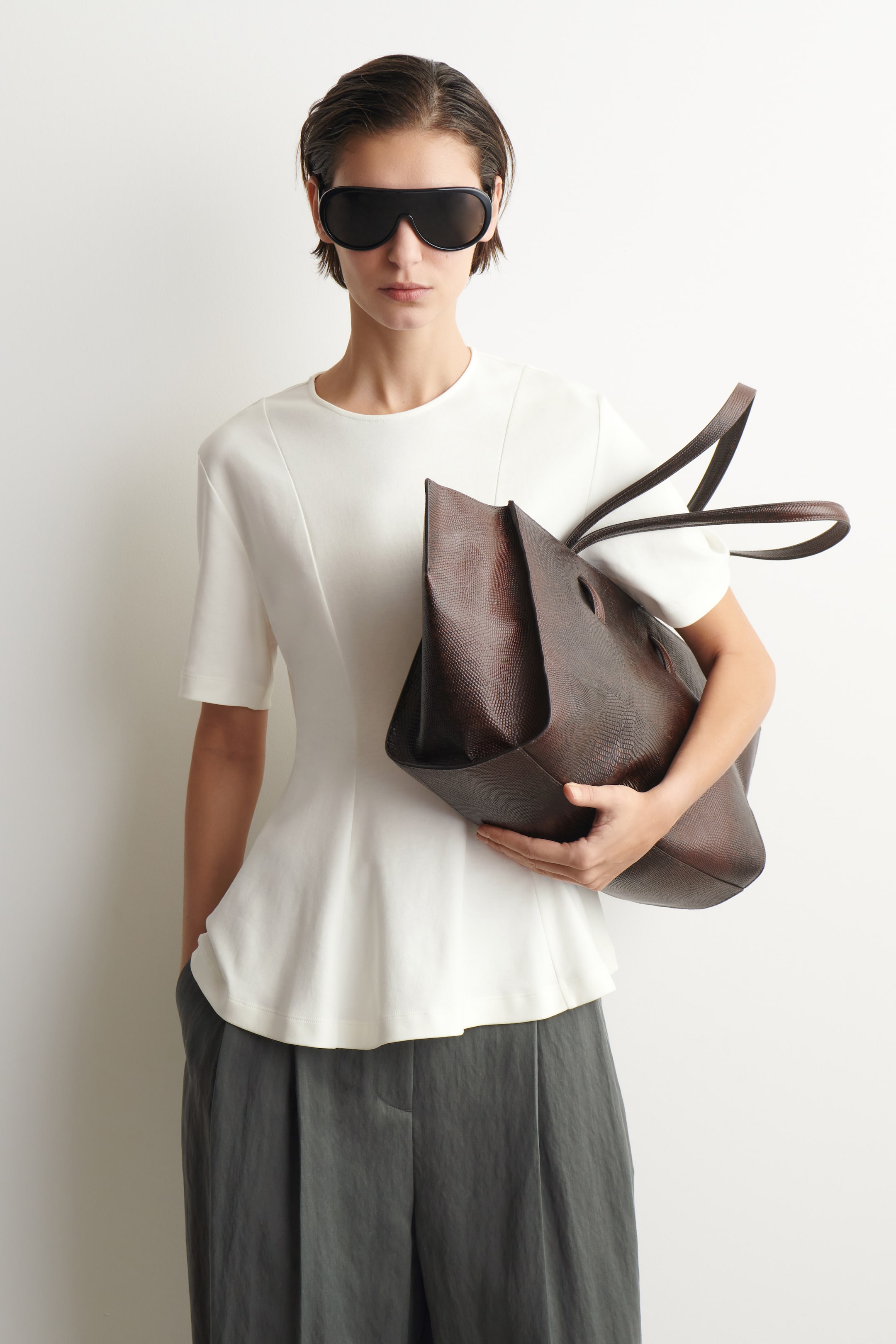 Agrandir l'image: Un sac fourre-tout marron foncé, avec une texture en relief imitant la peau de lézard, est tenu incliné vers le spectateur, mettant en valeur sa forme souple et non structurée ainsi que ses deux fines poignées.