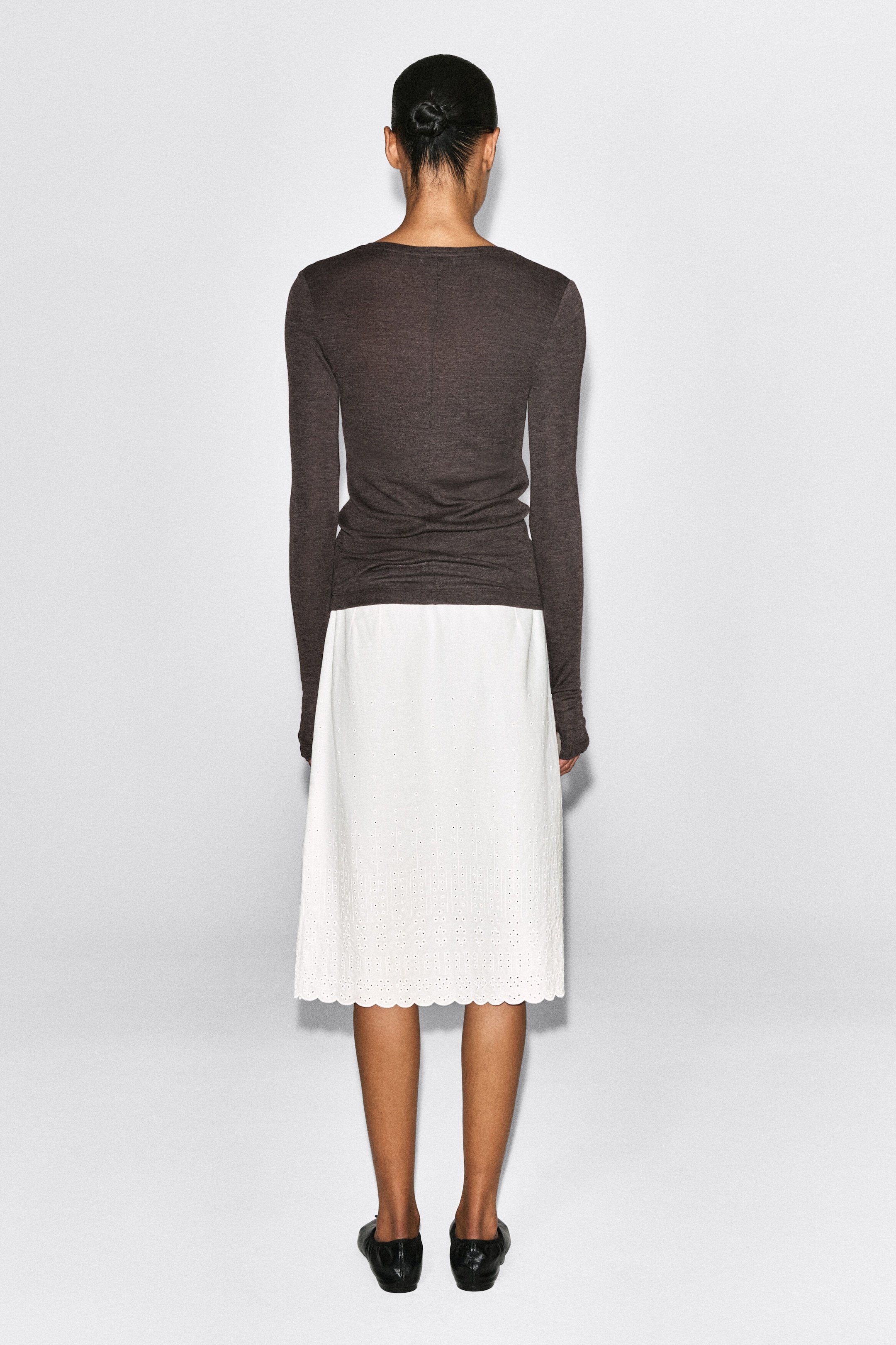 This slim merino wool top is shown from the back, featuring a close-fitting silhouette in a dark grey-brown fine knit with long sleeves. The top is layered over a white skirt, highlighting its straight hem and lightweight texture. This slim merino wool top is shown from the back, featuring a close-fitting silhouette in a dark grey-brown fine knit with long sleeves. The top is layered over a white skirt, highlighting its straight hem and lightweight texture.