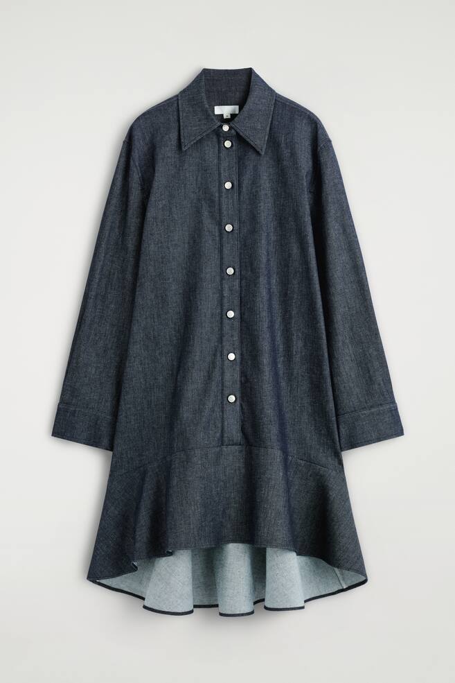 Un vestido camisero de denim azul oscuro está colgado, mostrado desde el FRENTE, con un cuello puntiagudo alargado, una parte frontal abotonada con botones blancos y mangas largas. El vestido está diseñado con una cintura caída y un dobladillo acampanado que es visiblemente más largo en la parte trasera. - 1