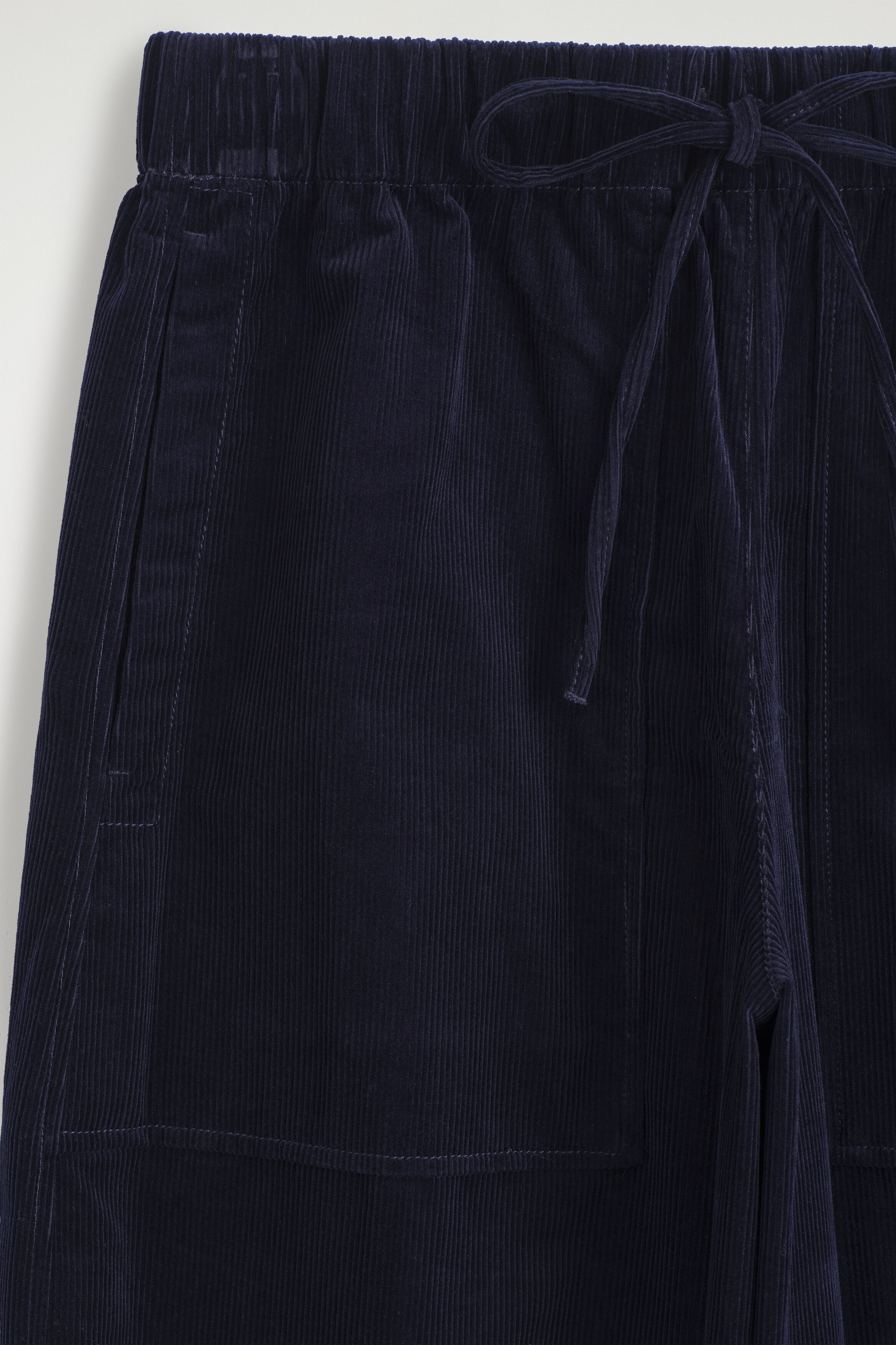 This close-up front view of the navy blue corduroy barrel-leg drawstring pants shows the elasticized waistband with a matching fabric drawstring tied in a bow. The fine-wale ribbed texture is visible, along with the stitching of a front patch pocket. This close-up front view of the navy blue corduroy barrel-leg drawstring pants shows the elasticized waistband with a matching fabric drawstring tied in a bow. The fine-wale ribbed texture is visible, along with the stitching of a front patch pocket.