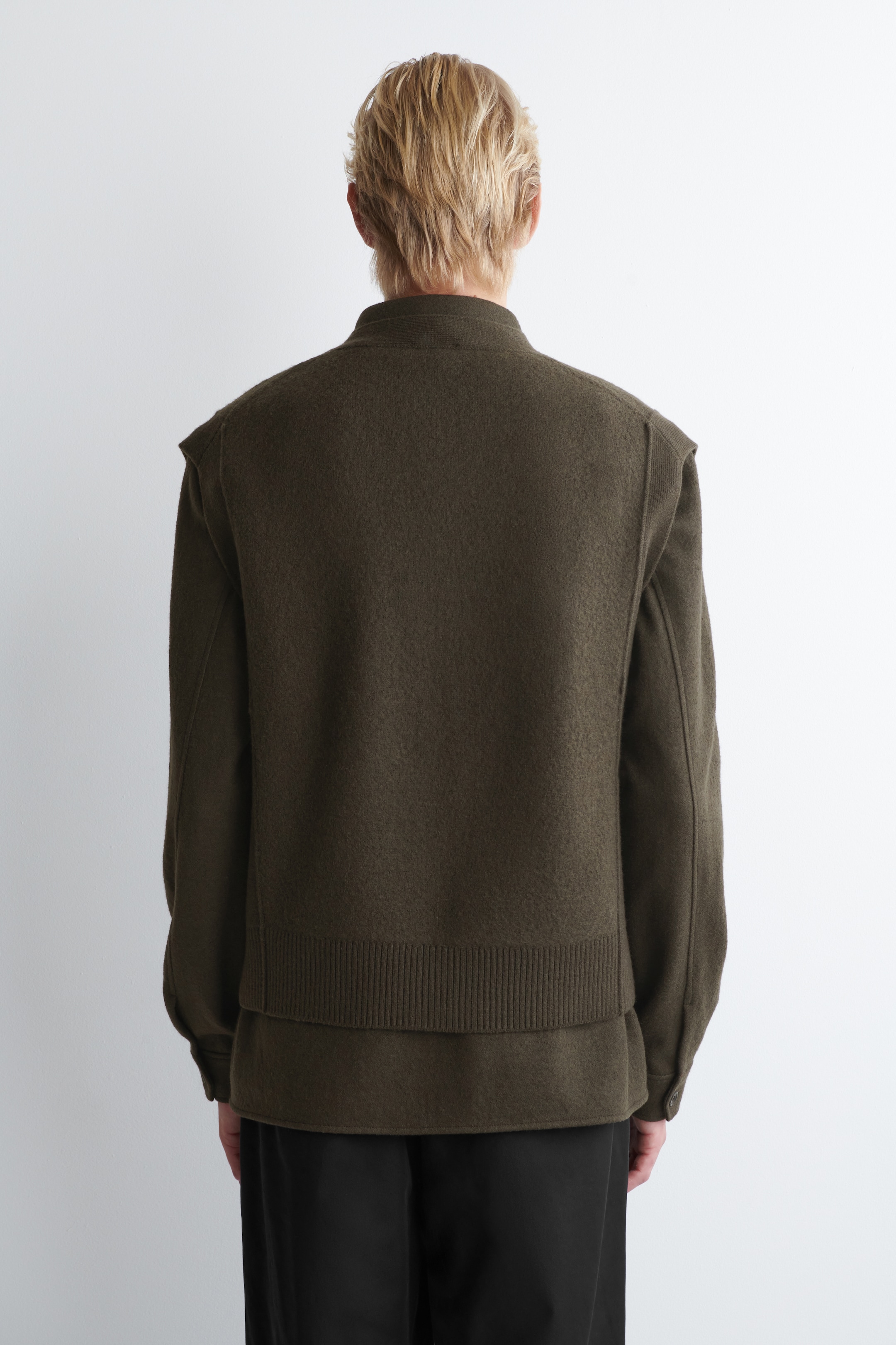 Agrandir l'image: Un gilet sans manches vert olive foncé est présenté de dos, superposé à une chemise à manches longues assortie. Le gilet présente un tissage texturé, un col haut et un ourlet large côtelé, tandis que les poignets boutonnés de la chemise se montrent aux poignets et que son ourlet s'étend sous le gilet.