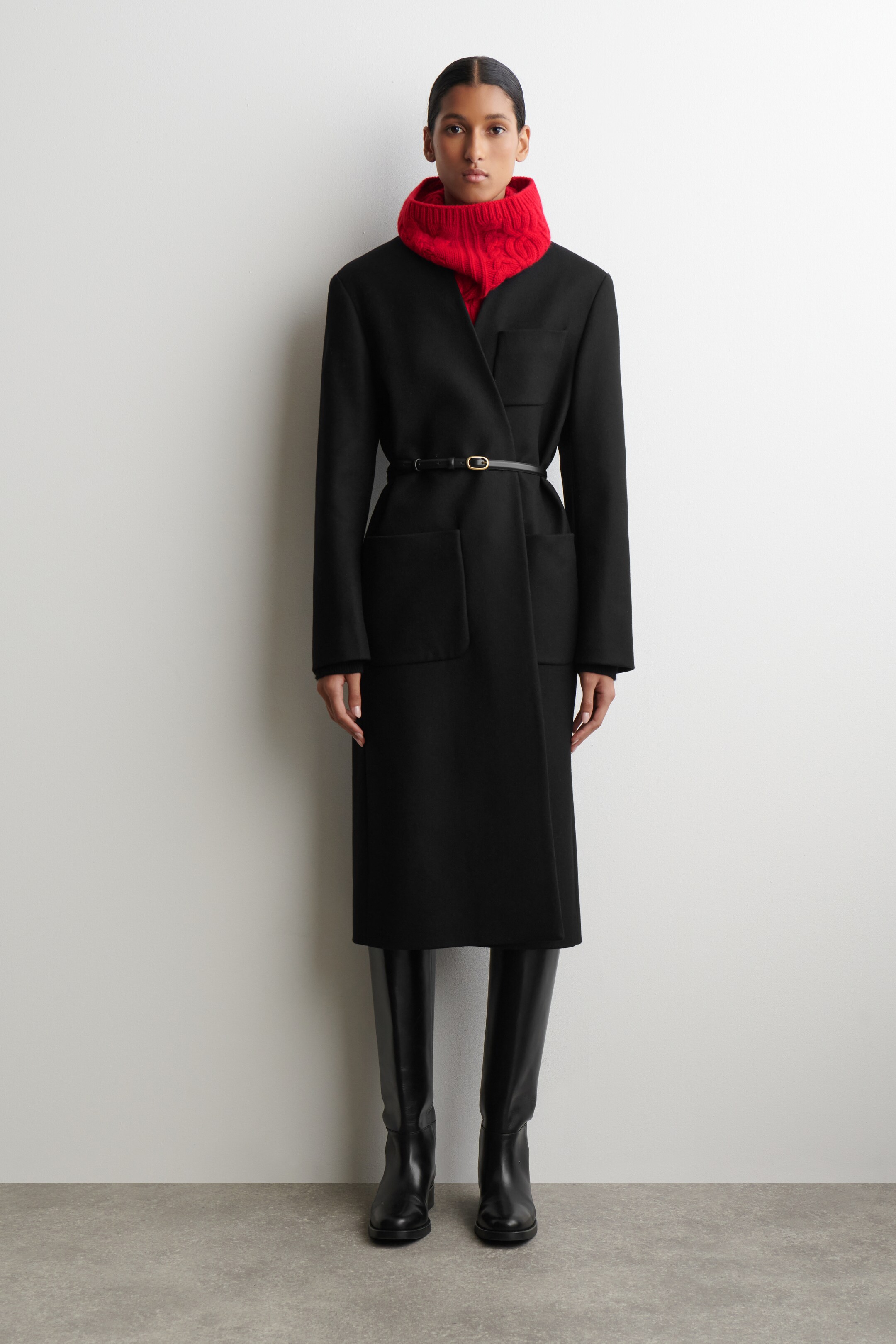 Agrandir l'image: Un long manteau noir est présenté face à l'avant, avec de larges revers en pointe et une fermeture de style portefeuille, comprenant deux grandes poches plaquées sur les hanches et une petite poche plaquée en haut à gauche. Une fine ceinture noire avec une boucle dorée serre la taille, et un col roulé rouge en maille torsadée est visible en dessous.
