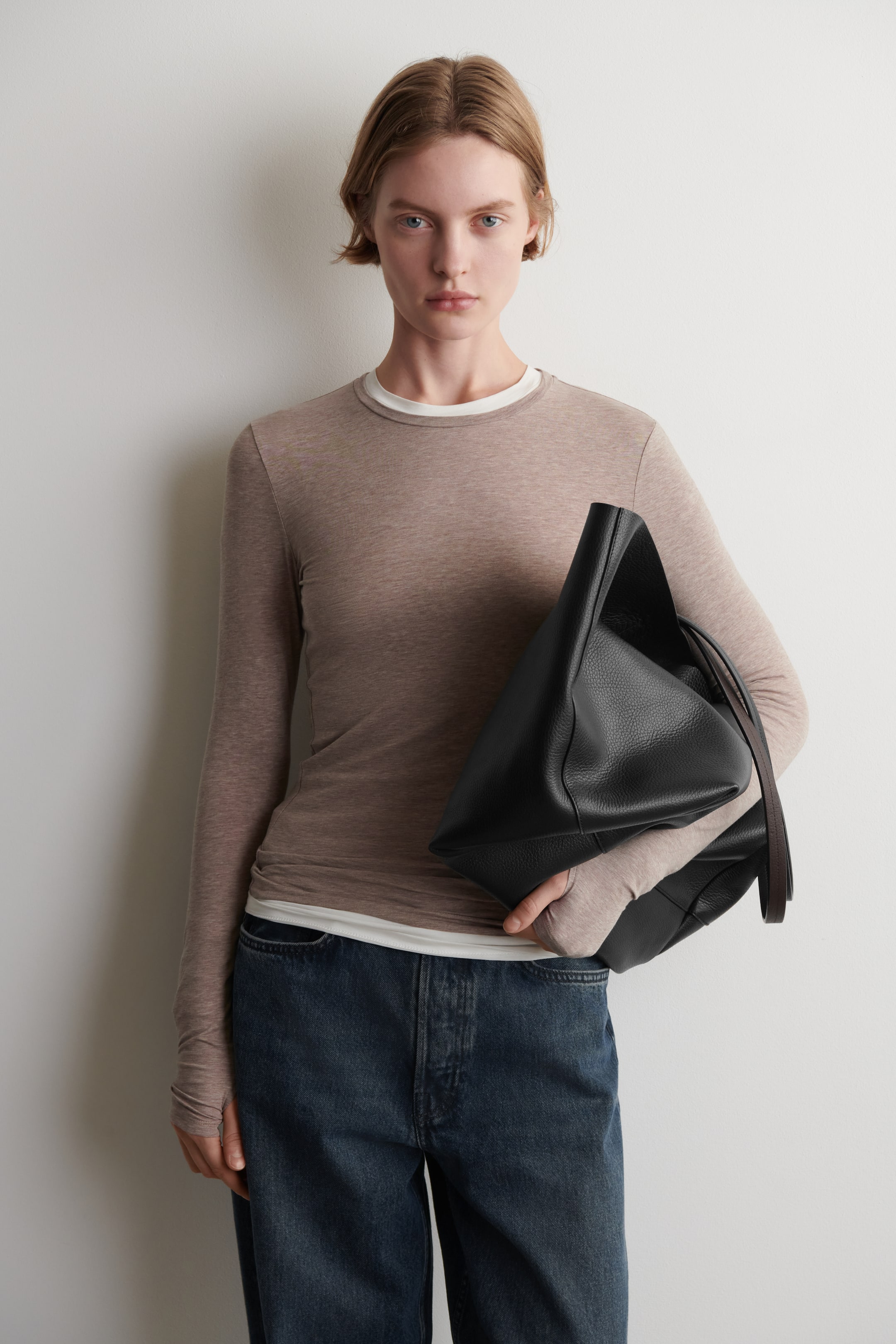 Agrandir l'image: Un sac en cuir grainé noir est tenu, ses panneaux avant et latéraux sont orientés vers l'avant, affichant sa forme douce et non structurée. Une sangle sombre pend du sac.