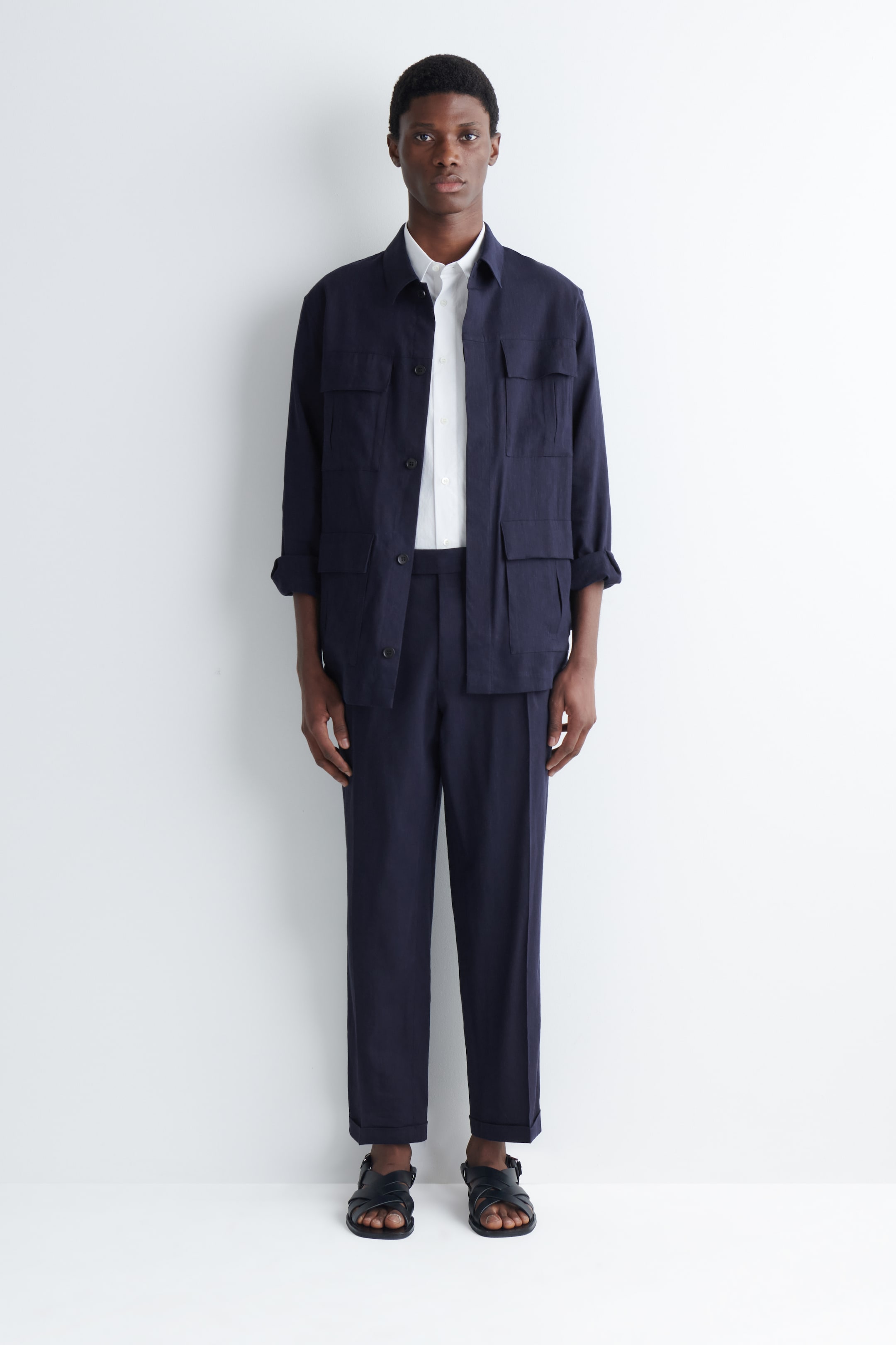 View larger image: A navy blue shacket and matching straight-leg trousers are showcased. The front-facing shacket features a spread collar, a button-front fastening, and four flap patch pockets, worn open over a white shirt, while the trousers fall to the ankles with cuffs, paired with black sandals.
