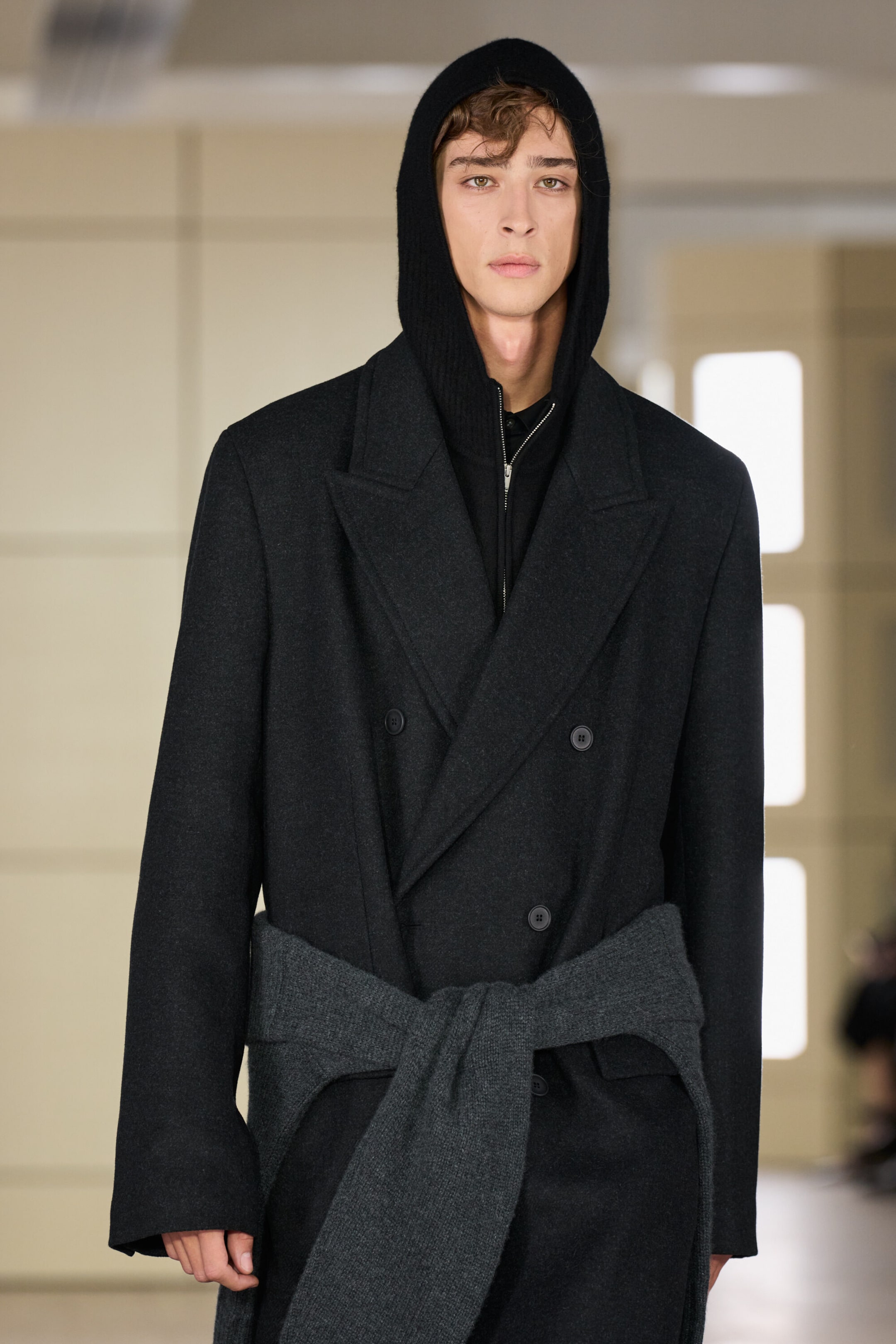 A front-facing view presents a black merino wool half-zip hoodie with the hood pulled up, layered beneath a structured charcoal double-breasted wool-blend long coat. The coat features wide lapels and button fastenings, with a grey knit jumper tied around the waist to complete the layered styling. A front-facing view presents a black merino wool half-zip hoodie with the hood pulled up, layered beneath a structured charcoal double-breasted wool-blend long coat. The coat features wide lapels and button fastenings, with a grey knit jumper tied around the waist to complete the layered styling.