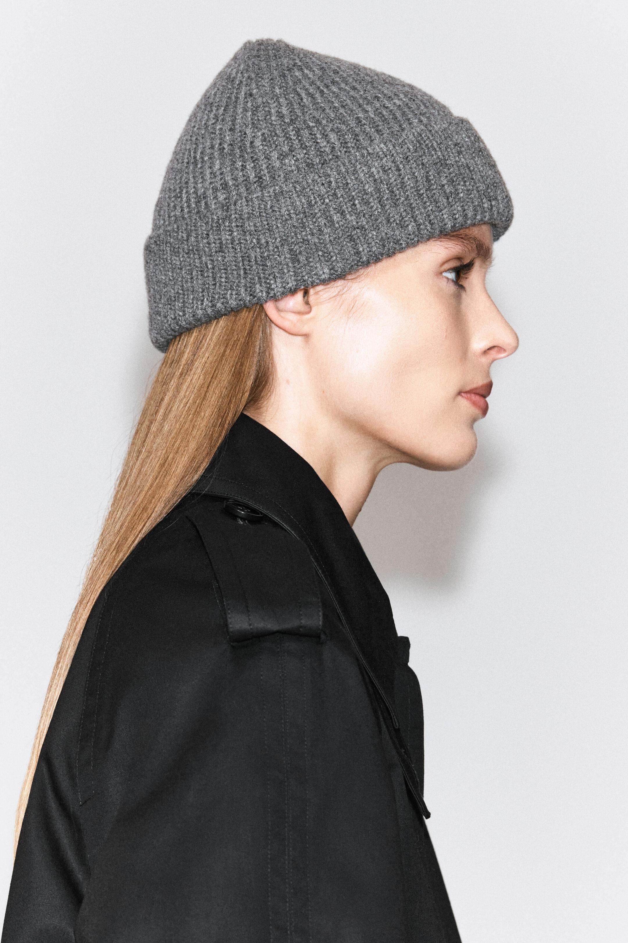 A gray ribbed knit beanie with a folded cuff is being worn by the model, who is shown in profile wearing a black trench coat. A gray ribbed knit beanie with a folded cuff is being worn by the model, who is shown in profile wearing a black trench coat.