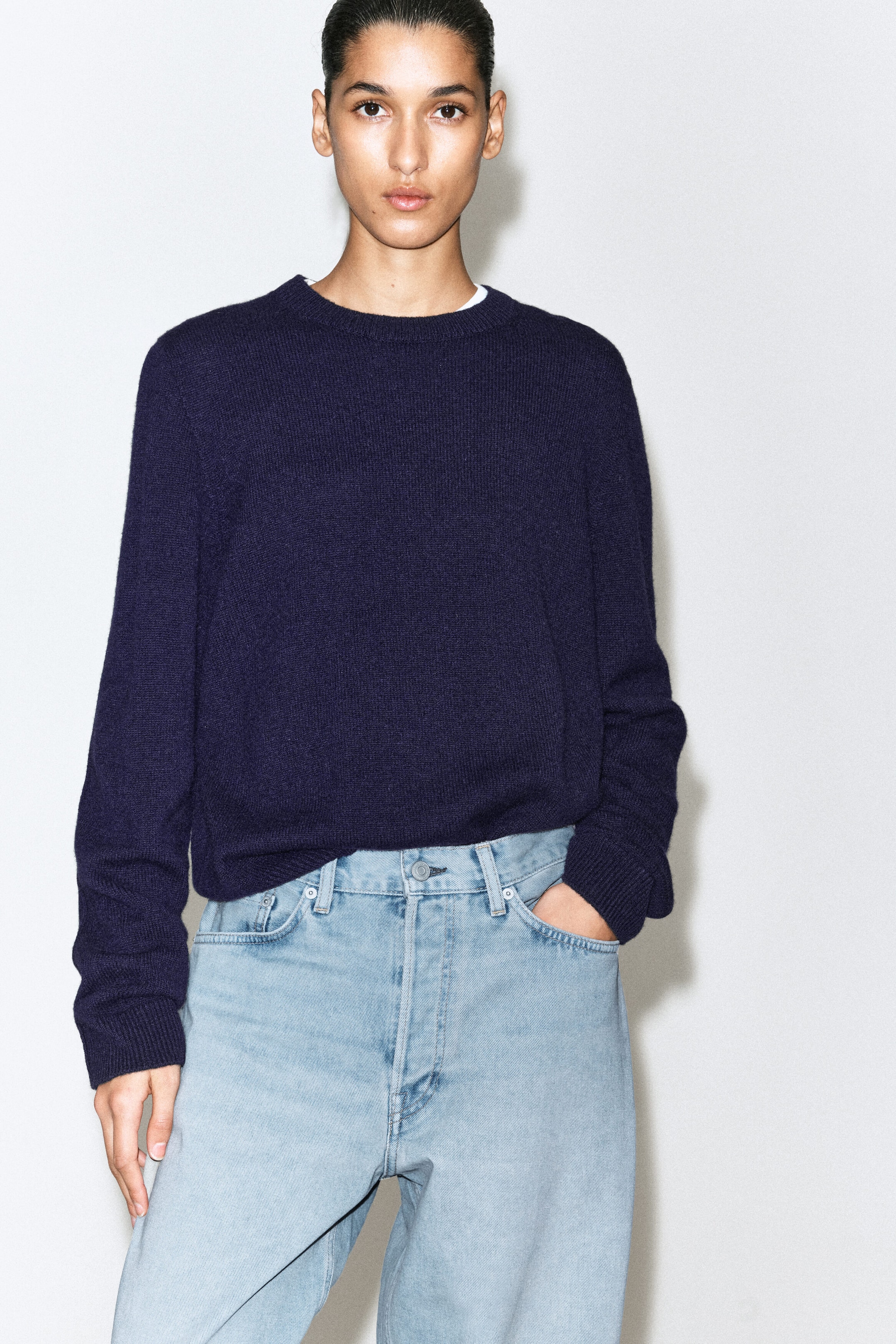 The Facade straight-leg jeans are shown in a light blue wash, featuring a structured waistband with belt loops and classic five-pocket styling at the top. A navy blue crew-neck sweater is resting over the waistband, with a hand slipping into the front pocket to showcase the relaxed, straight-leg fit.