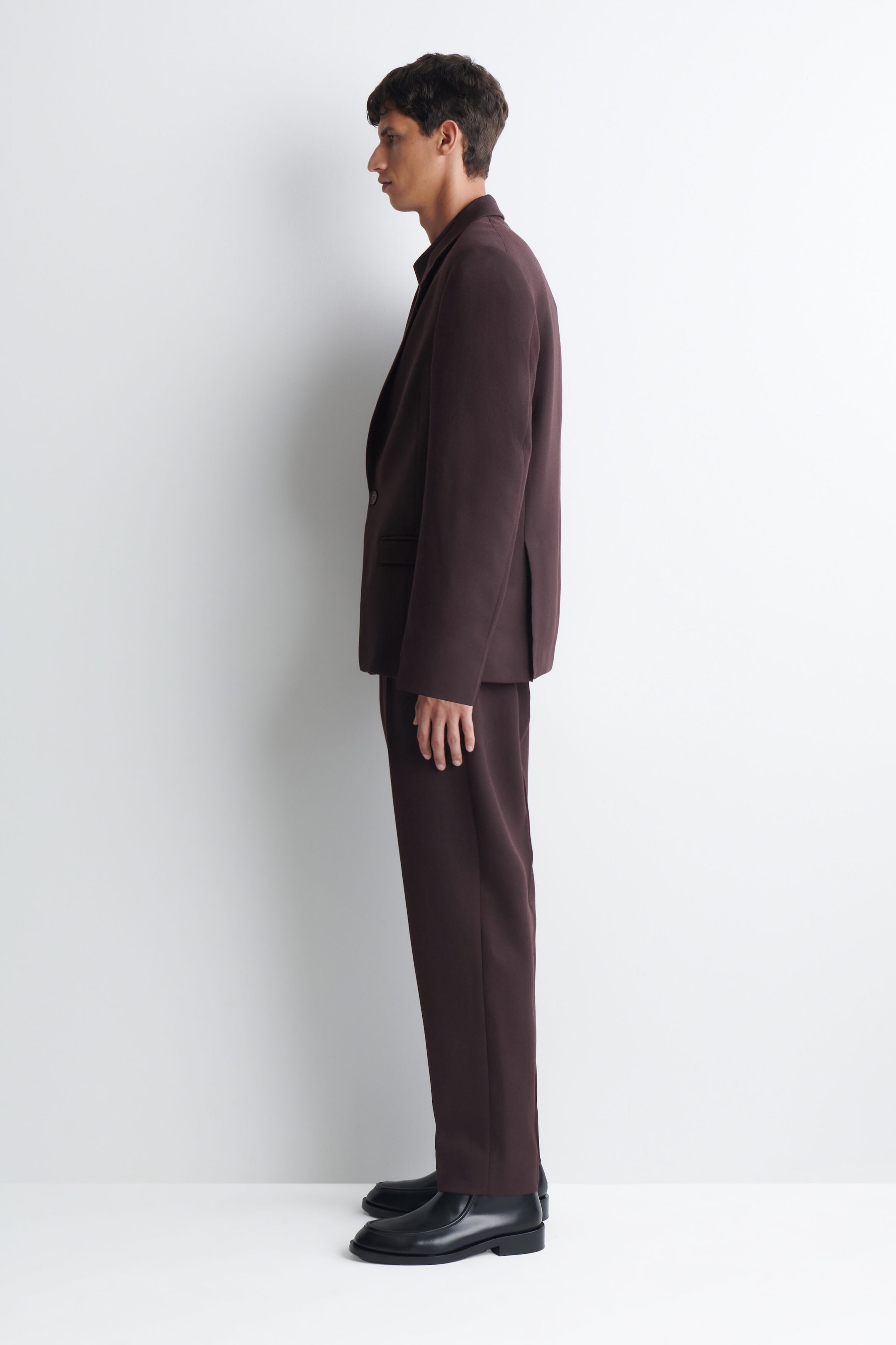 A dark brown suit, featuring a single-breasted blazer with a notched lapel and straight-leg trousers, is worn by the model. The blazer has one visible button, and the trousers extend to the model’s ankles, with black shoes completing the ensemble. A dark brown suit, featuring a single-breasted blazer with a notched lapel and straight-leg trousers, is worn by the model. The blazer has one visible button, and the trousers extend to the model’s ankles, with black shoes completing the ensemble.