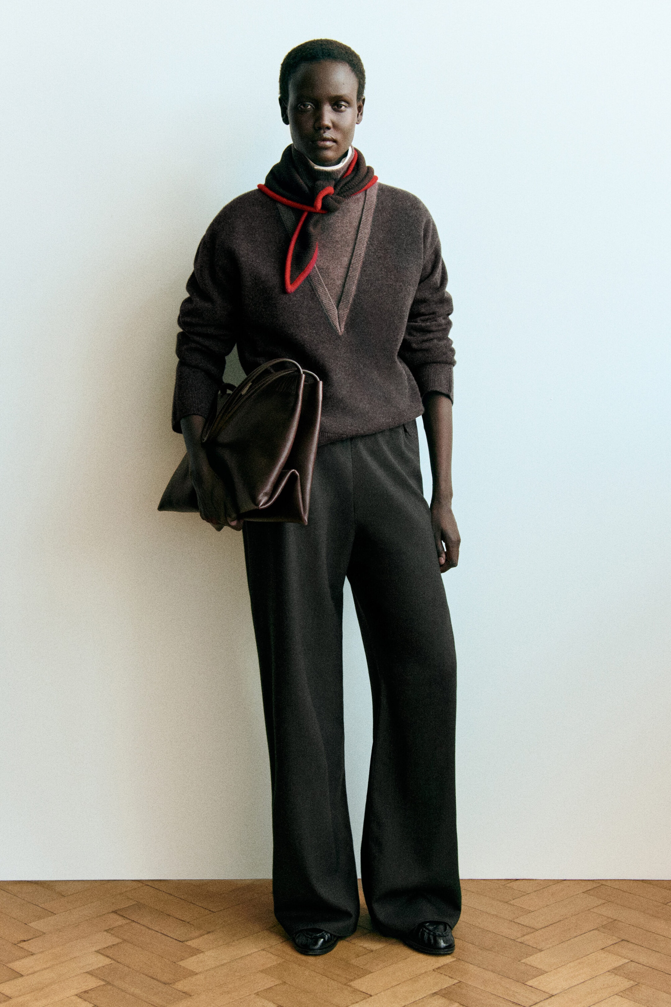The layered funnel-neck merino wool jumper in dark brown is shown from the front, featuring a deep V-neckline layered over a high collar with a contrasting red-trimmed tie. This relaxed-fit jumper has ribbed cuffs on the long sleeves and is styled with dark wide-leg trousers. The layered funnel-neck merino wool jumper in dark brown is shown from the front, featuring a deep V-neckline layered over a high collar with a contrasting red-trimmed tie. This relaxed-fit jumper has ribbed cuffs on the long sleeves and is styled with dark wide-leg trousers.
