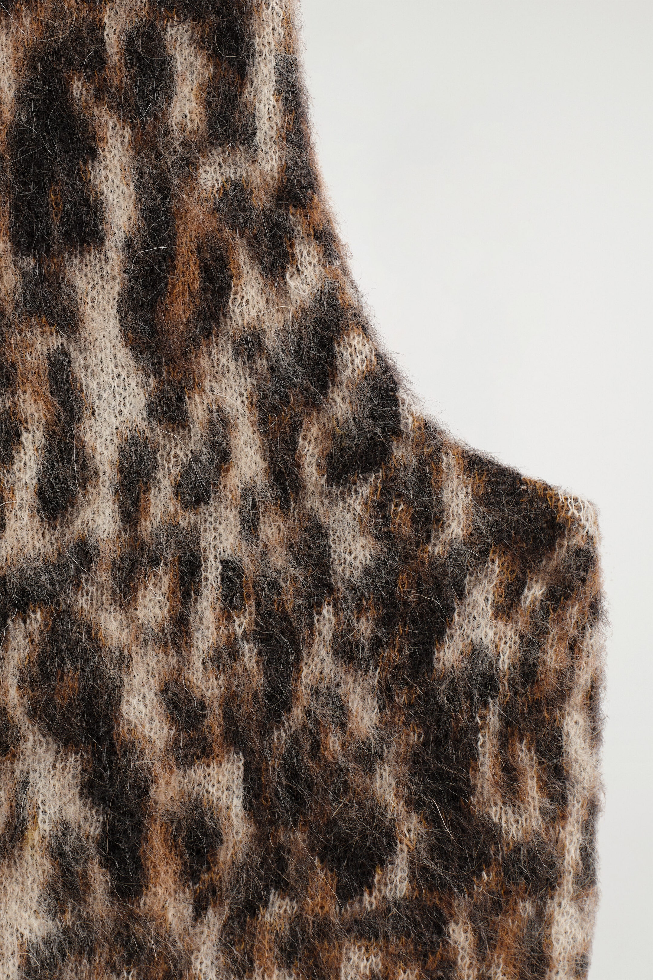 A close-up of a knit garment featuring a fuzzy leopard-print pattern in shades of brown, black, and cream. The garment displays a brushed, mohair-like texture with a curved edge outlining the armhole. A close-up of a knit garment featuring a fuzzy leopard-print pattern in shades of brown, black, and cream. The garment displays a brushed, mohair-like texture with a curved edge outlining the armhole.