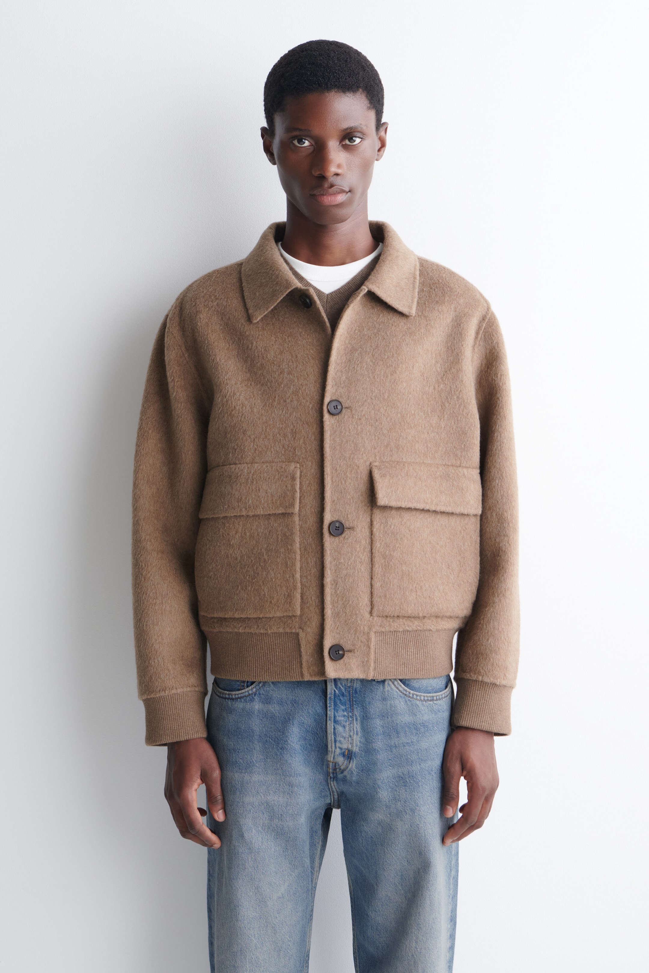 A double-faced blouson jacket in a textured camel-brown fabric is worn by the model, featuring a spread collar, front button closures, and two large flap pockets. The jacket is styled with a ribbed hem and cuffs and is layered over a brown knit sweater and blue jeans. A double-faced blouson jacket in a textured camel-brown fabric is worn by the model, featuring a spread collar, front button closures, and two large flap pockets. The jacket is styled with a ribbed hem and cuffs and is layered over a brown knit sweater and blue jeans.