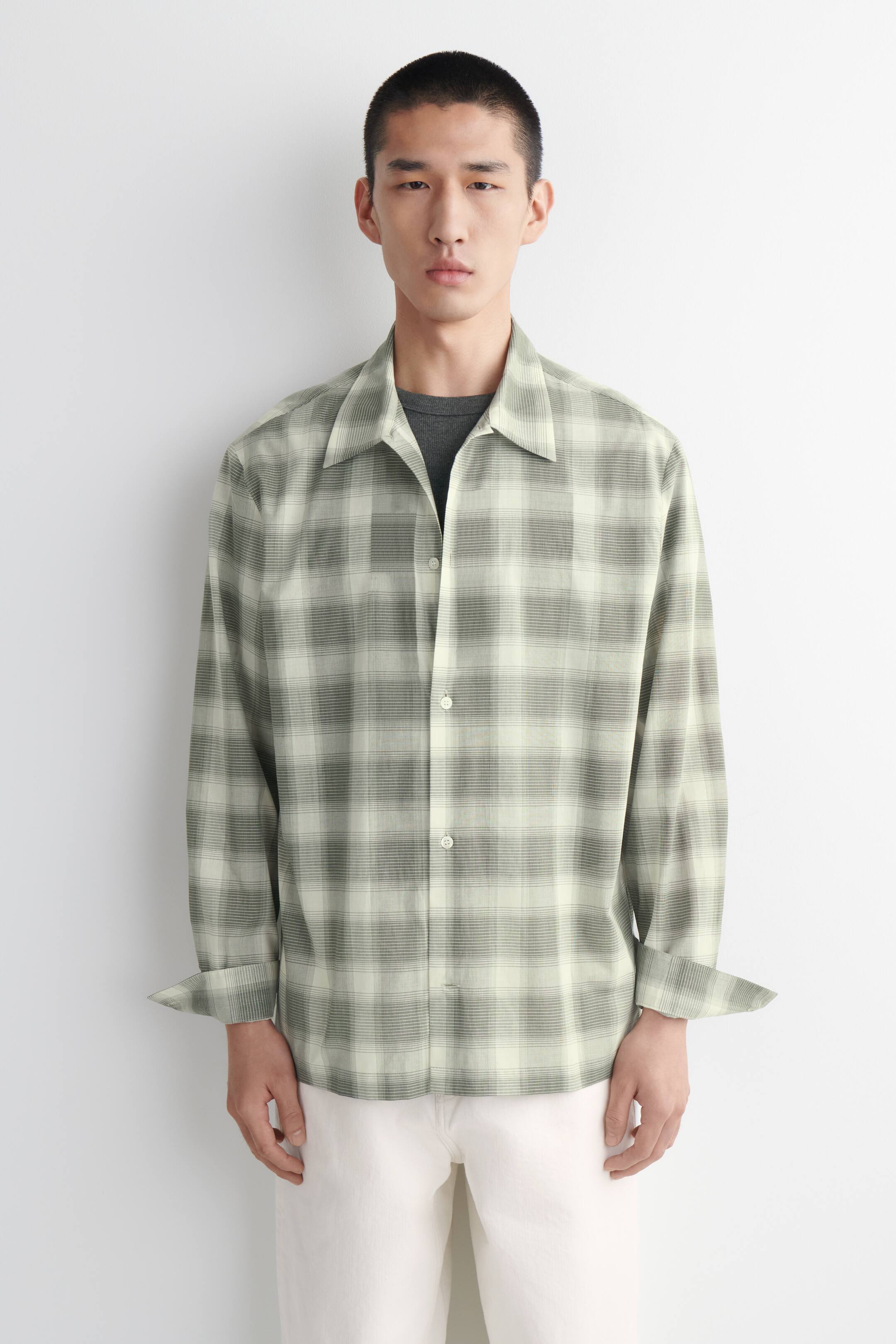 Größeres Bild anzeigen: Ein hellgrün und dunkelgrau verlaufendes Karohemd wird frontal präsentiert. Es verfügt über einen spitzen Kragen und hellfarbige Knöpfe entlang der Vorderseite, und die langen Ärmel sind an den Manschetten hochgekrempelt. Das Hemd wird offen über einem dunkelgrauen T-Shirt getragen.