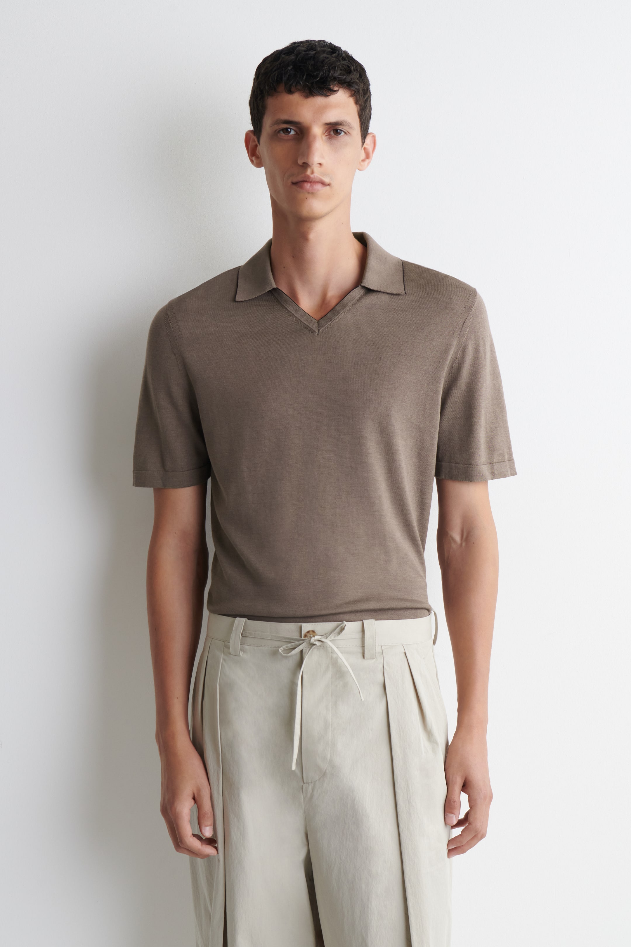 Größeres Bild ansehen: Ein braunes Strick-Poloshirt wird von vorne präsentiert, mit einem Kentkragen, einem V-Ausschnitt und kurzen Ärmeln. Es wird in eine hellfarbene, plissierte Hose gesteckt getragen.