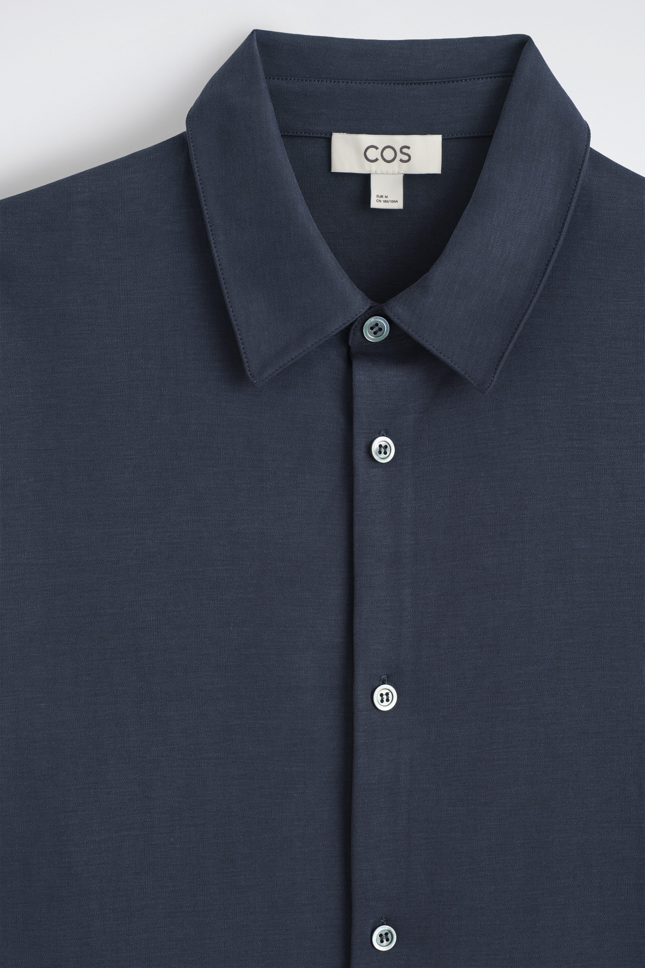 A close-up front view of the fluid interlock jersey shirt showing its dark navy blue fabric, pointed collar, and neat button placket. The shirt features light-colored buttons and a visible white brand label sewn inside the collar. A close-up front view of the fluid interlock jersey shirt showing its dark navy blue fabric, pointed collar, and neat button placket. The shirt features light-colored buttons and a visible white brand label sewn inside the collar.
