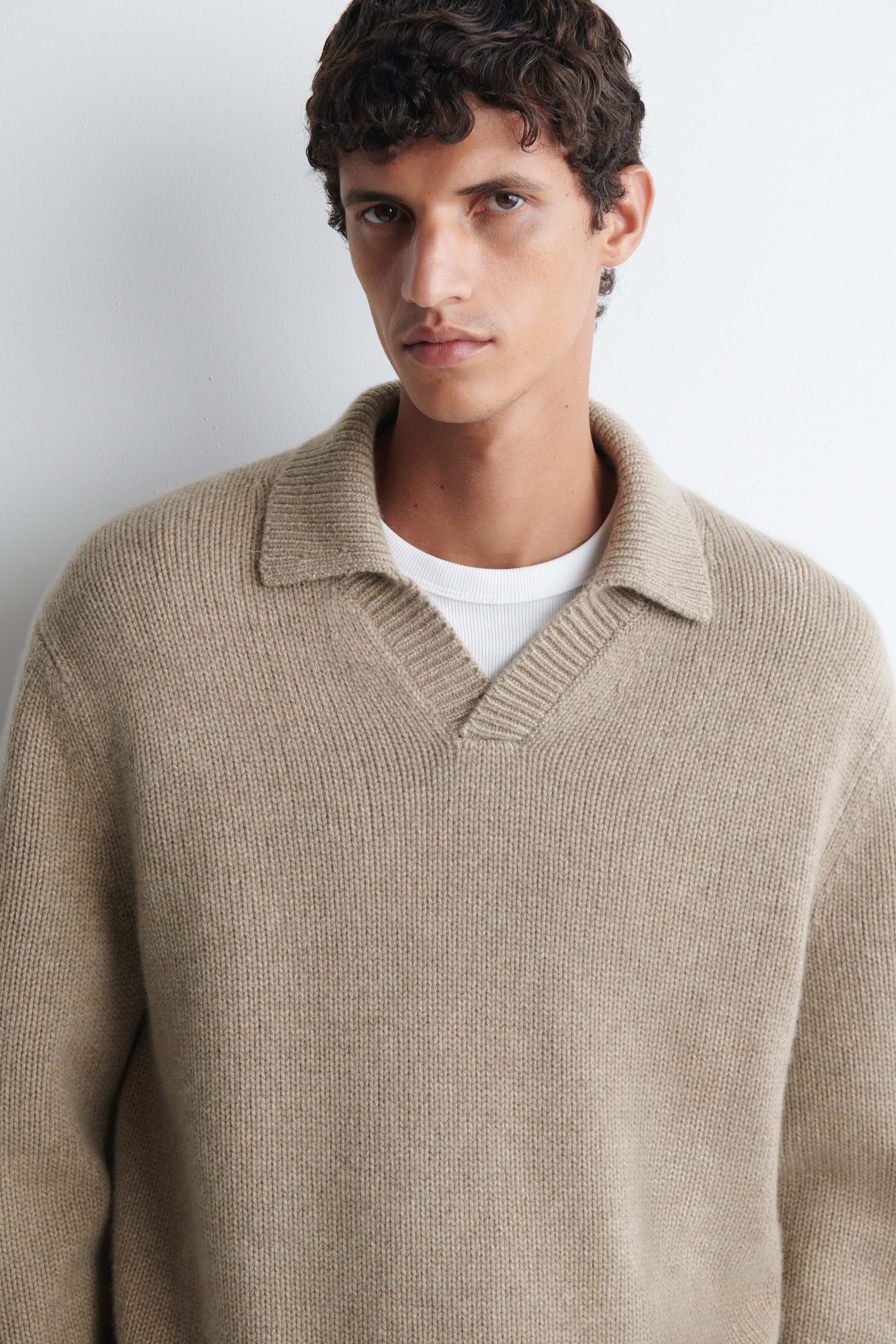 A beige knit polo sweater featuring a ribbed texture, a classic collar, and an open V-neck is being worn by the model. The sweater is layered over a white crewneck t-shirt. A beige knit polo sweater featuring a ribbed texture, a classic collar, and an open V-neck is being worn by the model. The sweater is layered over a white crewneck t-shirt.