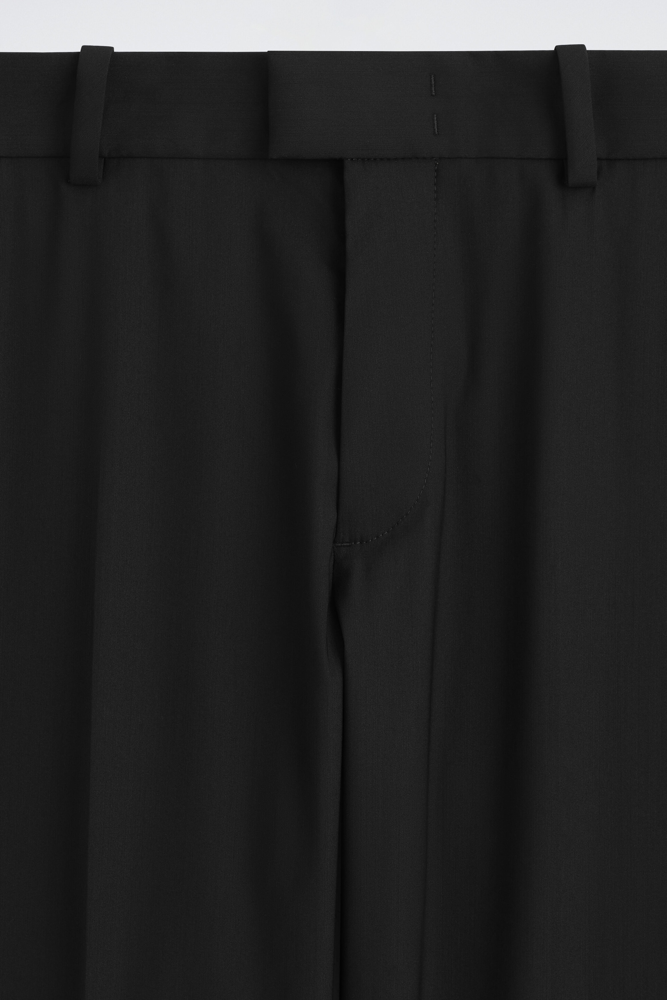 Spodnie o prostym kroju, slim fit, z domieszką wełny, prezentowane są w zbliżeniu z przodu, obejmującym pas i rozporek. Projekt wyróżnia się usztywnionym paskiem z szlufkami na pasek, przedłużonym zapięciem na patkę oraz ukrytym rozporkiem na zamek błyskawiczny z gładkiej czarnej tkaniny.