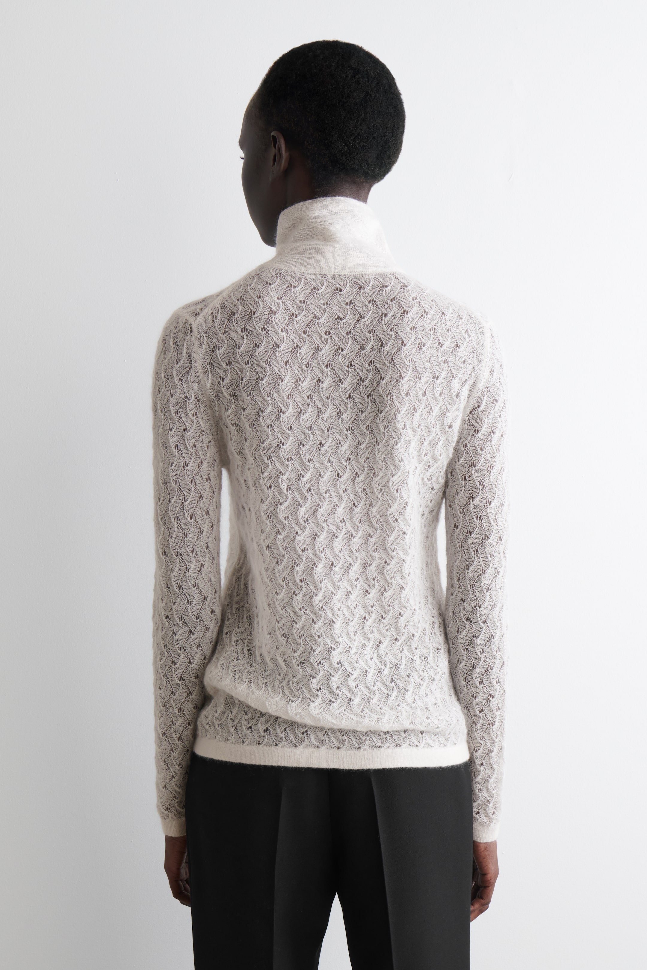 Grösseres Bild ansehen: Ein hellbeiges, langärmliges Oberteil mit schlanker Passform wird von hinten gezeigt, mit einem subtilen Pointelle-Strickmuster, das einen durchscheinenden Effekt über den Körper und die Ärmel erzeugt. Es ist mit einem gerippten Trichterkragen, gerippten Bündchen und einem gerippten Saum gestaltet.