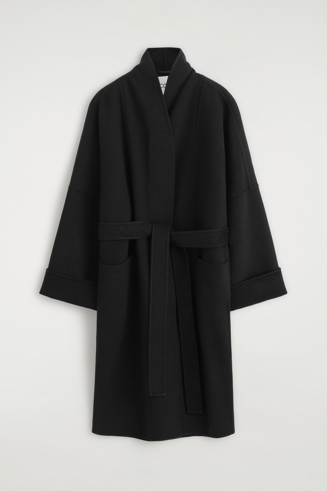 Ce manteau châle en laine double face oversize noir est suspendu de face, mettant en valeur une silhouette décontractée avec un grand col châle et des manches larges retroussées. Le manteau est doté d’une ceinture assortie à nouer à la taille et de deux poches plaquées sur le devant.