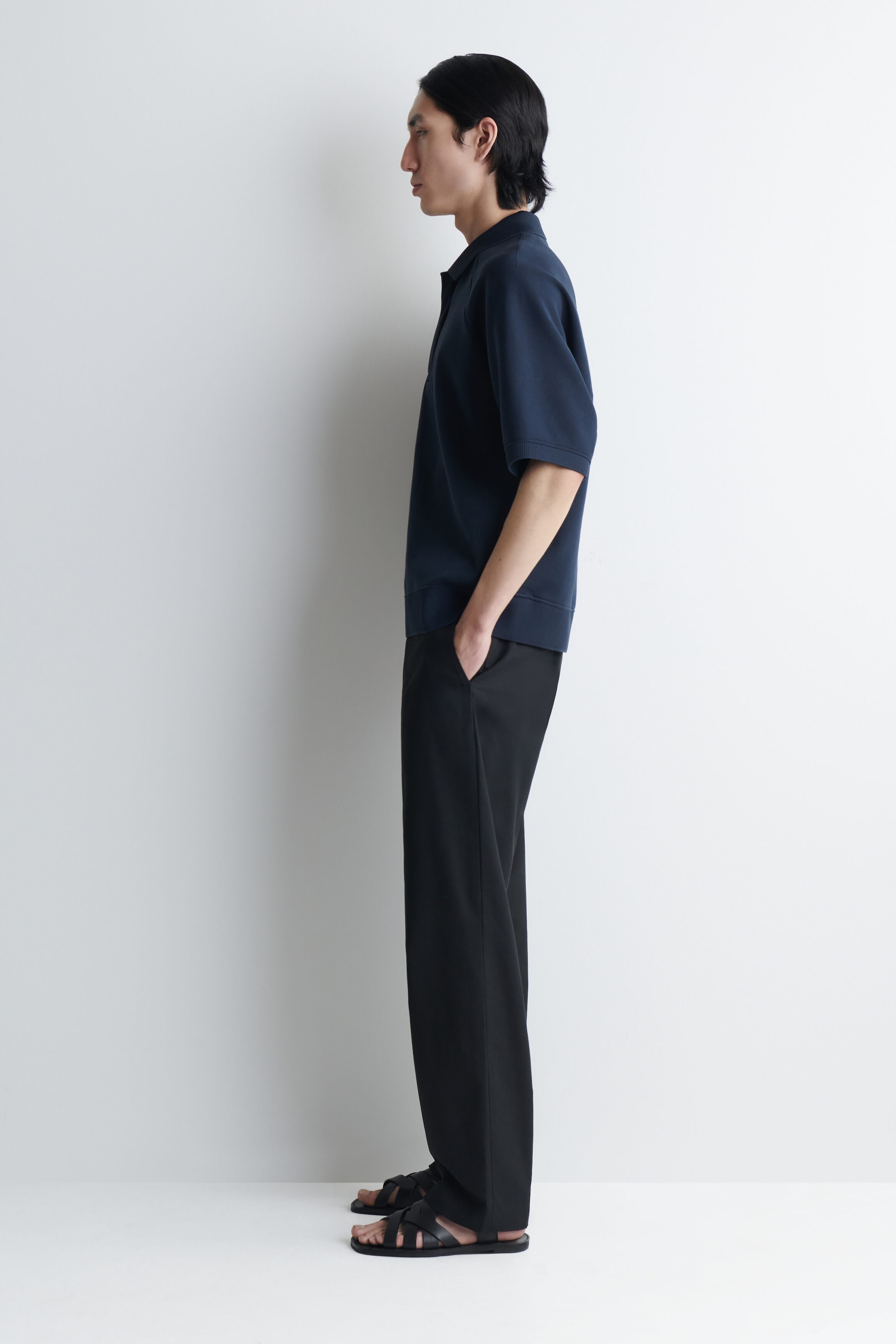 A navy blue scuba jersey polo shirt is being worn by the model, shown in side profile. The short-sleeved shirt features a structured drape, a classic collar, and a ribbed hem, and is styled with black trousers. A navy blue scuba jersey polo shirt is being worn by the model, shown in side profile. The short-sleeved shirt features a structured drape, a classic collar, and a ribbed hem, and is styled with black trousers.