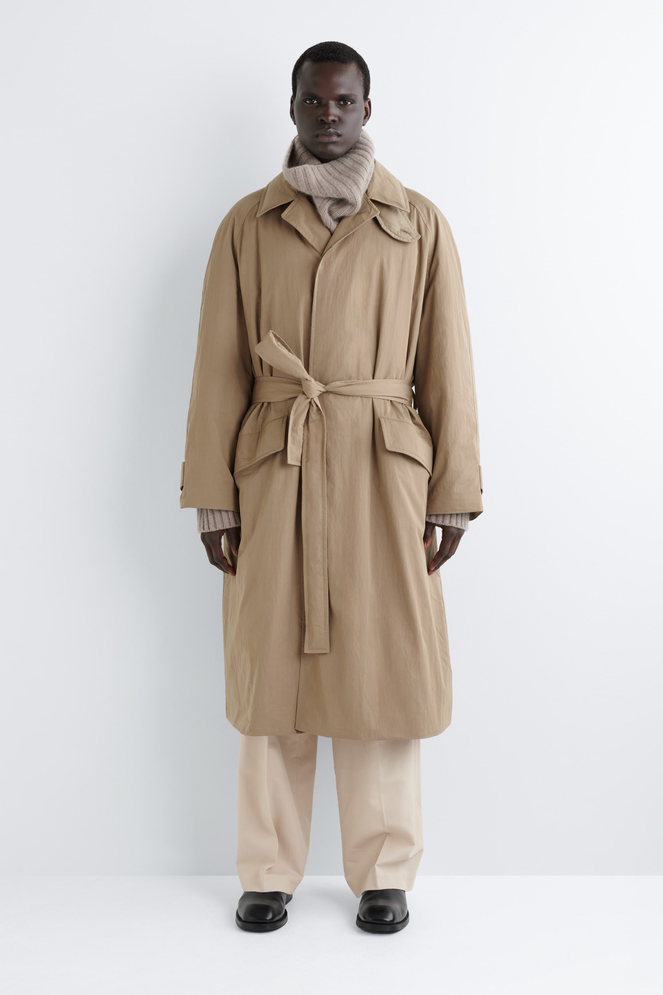 Visualizza immagine più grande: Un lungo cappotto beige semplice è presentato frontalmente, con un colletto a punta con una chiusura a levetta visibile e una chiusura con bottoni parzialmente visibile. Il cappotto si stringe in vita con una cintura in tessuto annodata a nodo, e due grandi tasche applicate sono visibili sotto la cintura. Un maglione a maglia di colore chiaro a coste è stratificato al collo e alle maniche.