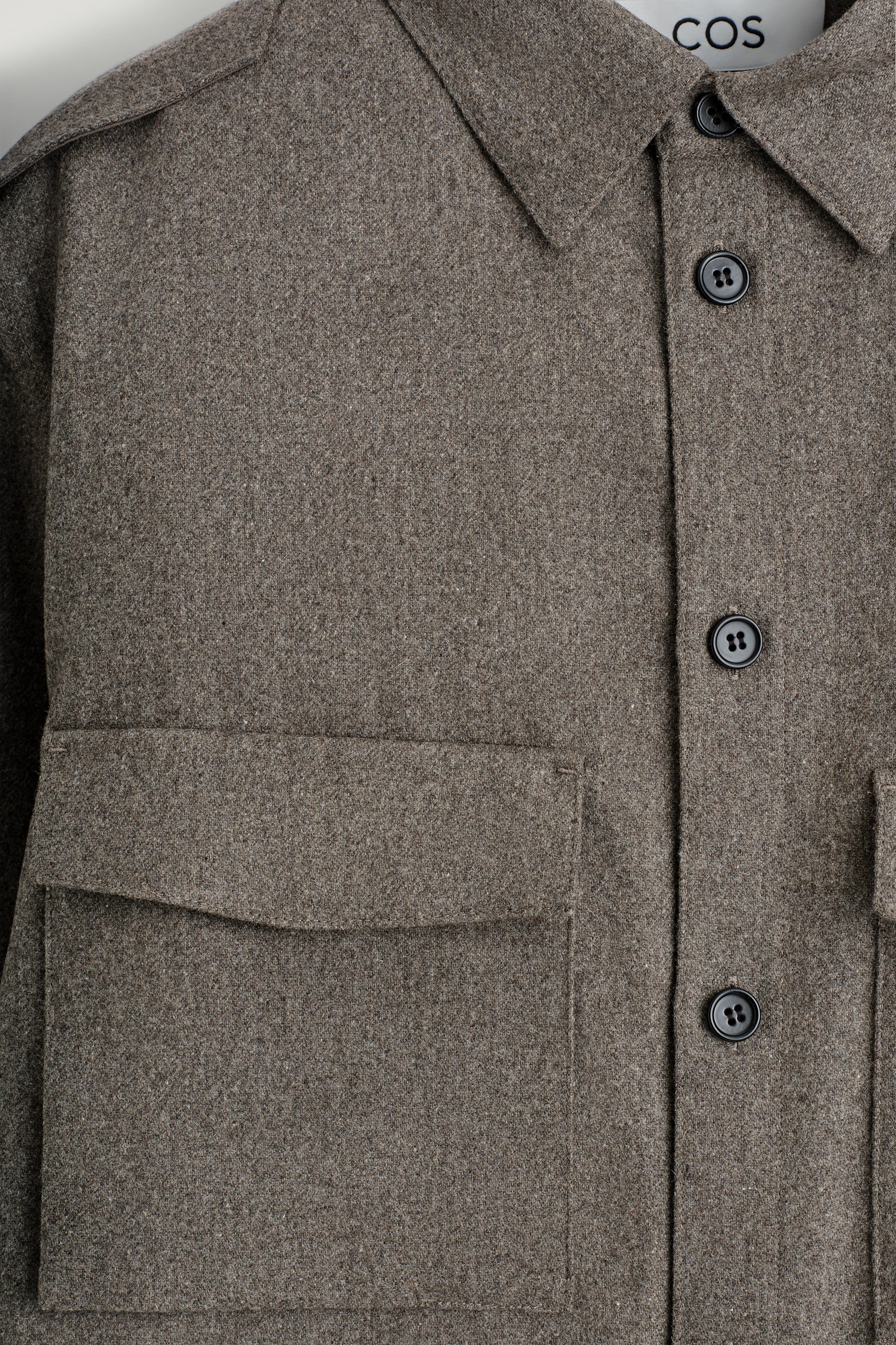 View larger image: A close-up view is showing the front of a heathered brown utility shirt. It is featuring a classic collar, black button closure down the front, a patch pocket with a flap on the chest, and epaulette details on the shoulders.