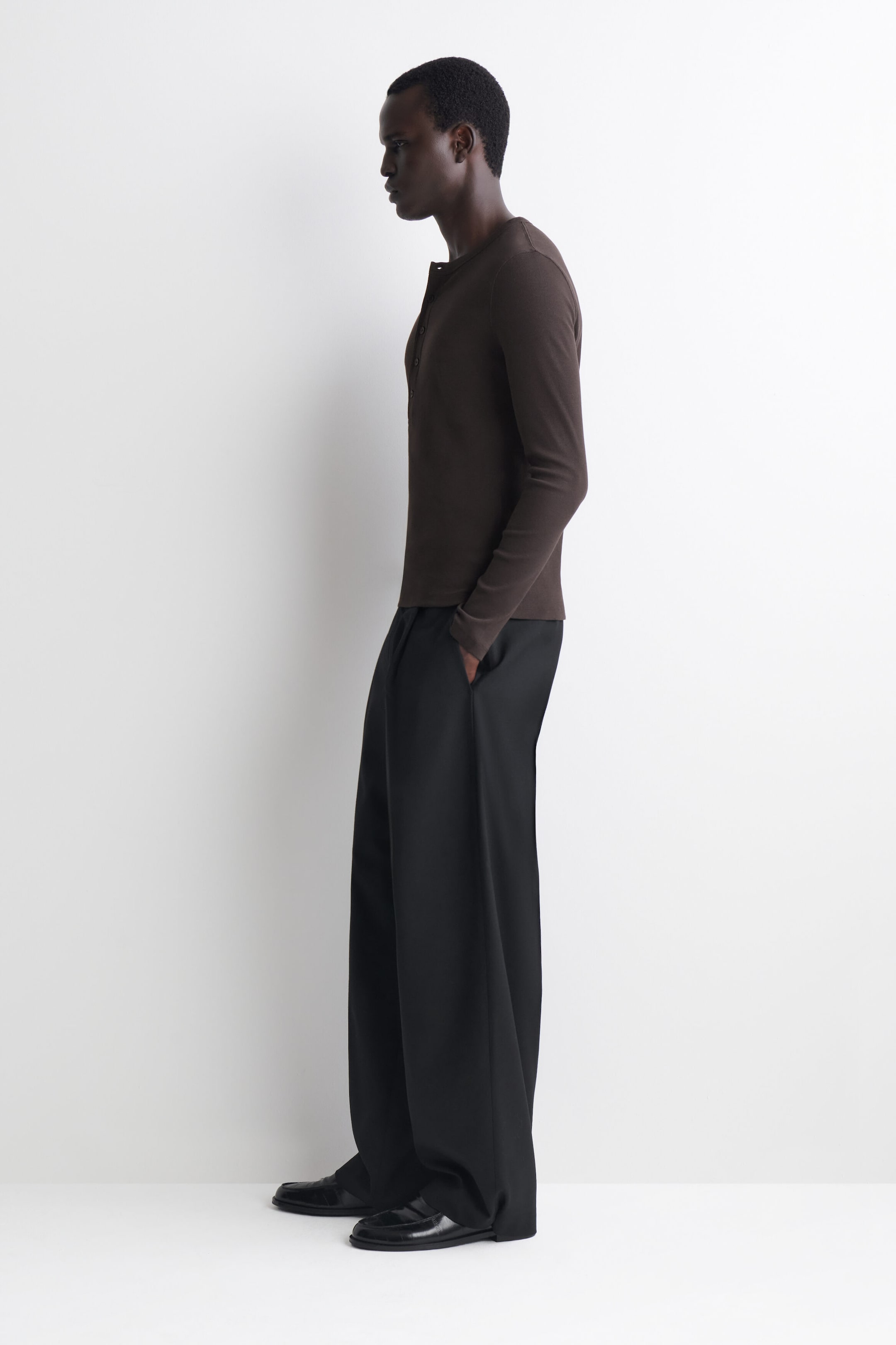 Obcisły bawełniany longsleeve Henley z drobnym prążkiem w ciemnobrązowym kolorze jest prezentowany z lewej strony, podkreślając dopasowany fason i delikatną prążkowaną fakturę. Ma częściowo rozpięty dekolt z listwą guzikową i jest noszony luźno na wierzchu, na szerokich czarnych spodniach z prostą, szeroką nogawką. Obcisły bawełniany longsleeve Henley z drobnym prążkiem w ciemnobrązowym kolorze jest prezentowany z lewej strony, podkreślając dopasowany fason i delikatną prążkowaną fakturę. Ma częściowo rozpięty dekolt z listwą guzikową i jest noszony luźno na wierzchu, na szerokich czarnych spodniach z prostą, szeroką nogawką.