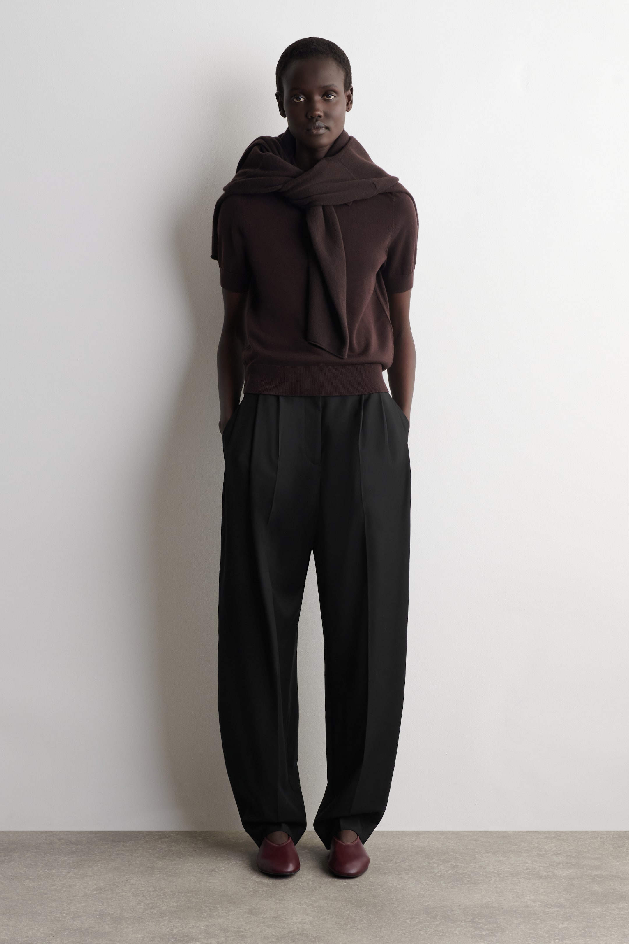 Visa större bild: En mörkbrun kortärmad stickad topp med ribbad nederkant bärs, med en matchande mörkbrun stickad tröja draperad över axlarna och knuten framtill. Svarta byxor med vida ben och frontveck bärs, och mörkröda platta skor syns på fötterna.