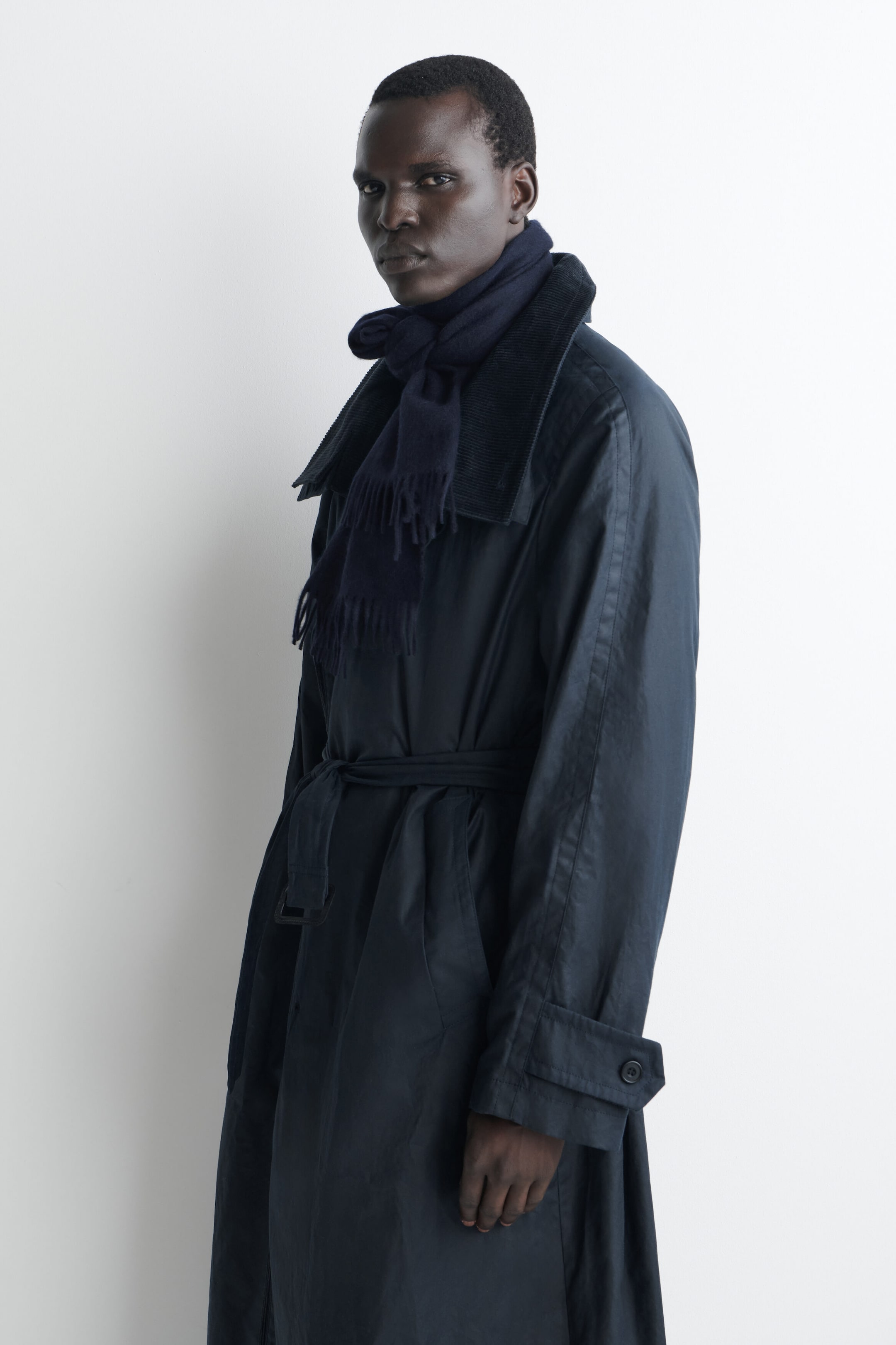 A dark navy blue fringed cashmere scarf is worn knotted around the neck, with its fringed ends hanging down the front over a dark trench coat. The product is shown on a model facing three-quarters left, highlighting the scarf’s soft texture and structured drape. A dark navy blue fringed cashmere scarf is worn knotted around the neck, with its fringed ends hanging down the front over a dark trench coat. The product is shown on a model facing three-quarters left, highlighting the scarf’s soft texture and structured drape.