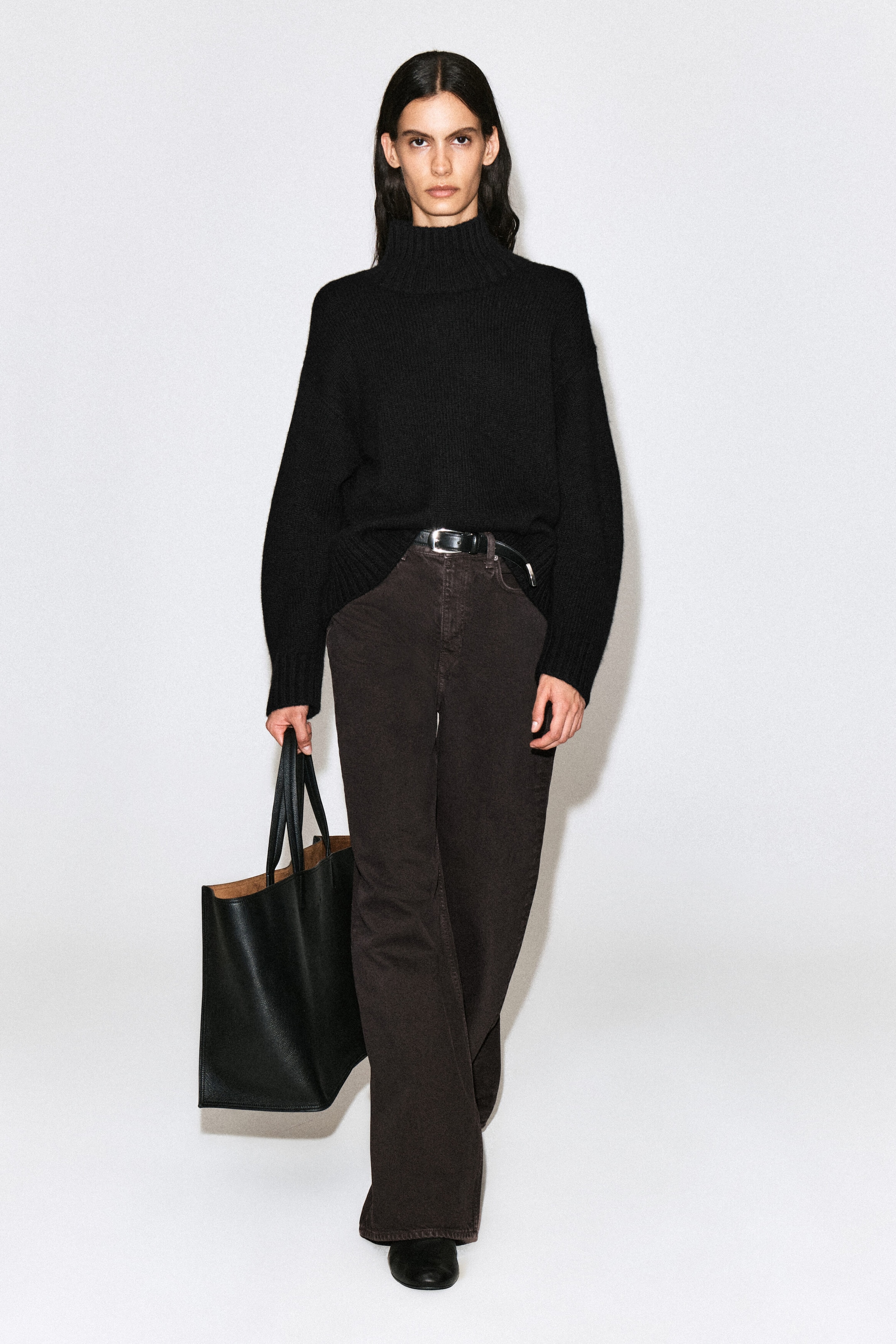 The model is wearing dark brown wide-leg jeans, styled with a black leather belt and a black turtleneck sweater. She is also carrying a large black leather tote bag in one hand. The model is wearing dark brown wide-leg jeans, styled with a black leather belt and a black turtleneck sweater. She is also carrying a large black leather tote bag in one hand.