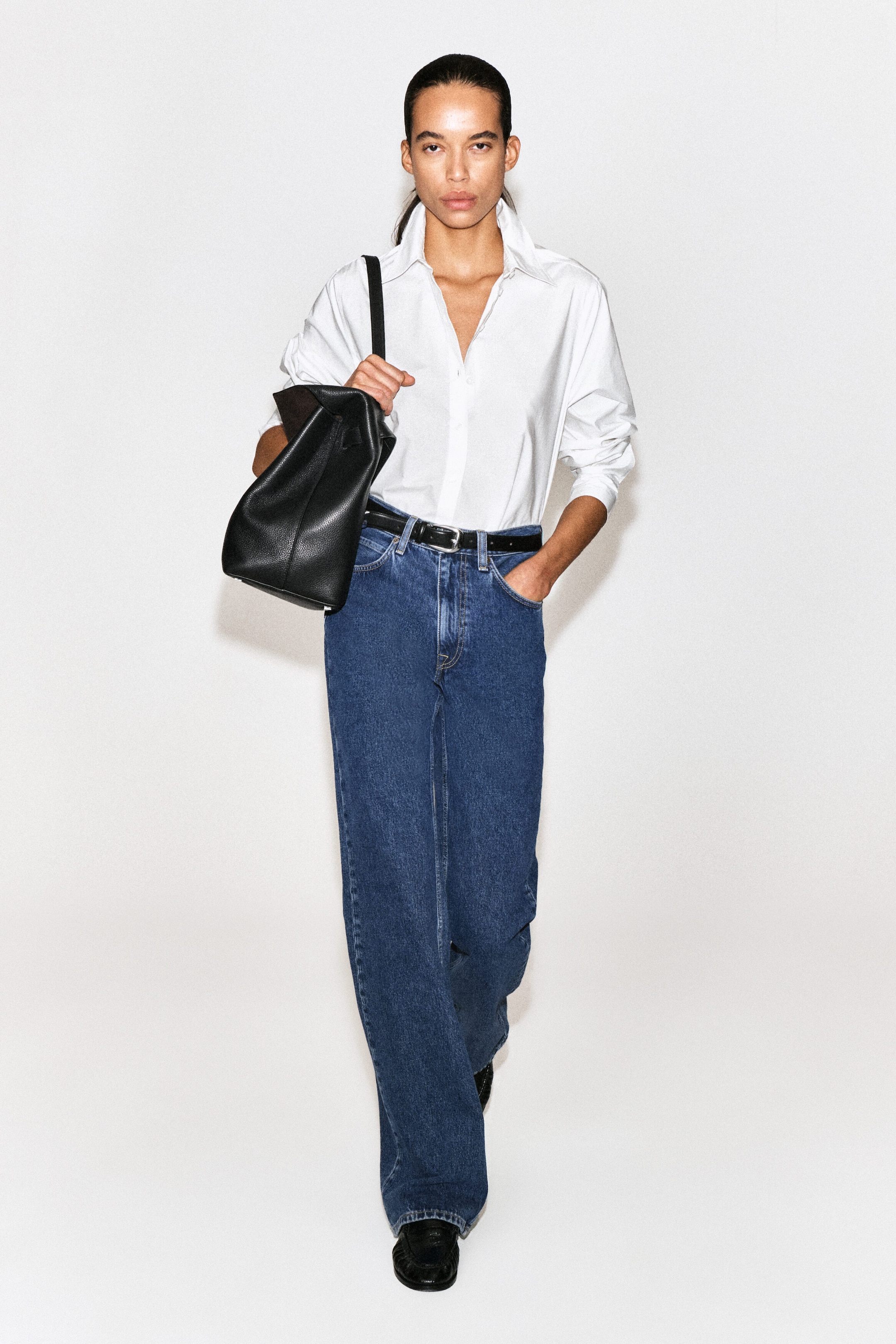 A classic white cotton poplin shirt is shown from the front, featuring an open pointed collar and long sleeves pushed up to the forearms. The shirt is styled tucked into high-waisted blue jeans, highlighting its relaxed silhouette. A classic white cotton poplin shirt is shown from the front, featuring an open pointed collar and long sleeves pushed up to the forearms. The shirt is styled tucked into high-waisted blue jeans, highlighting its relaxed silhouette.