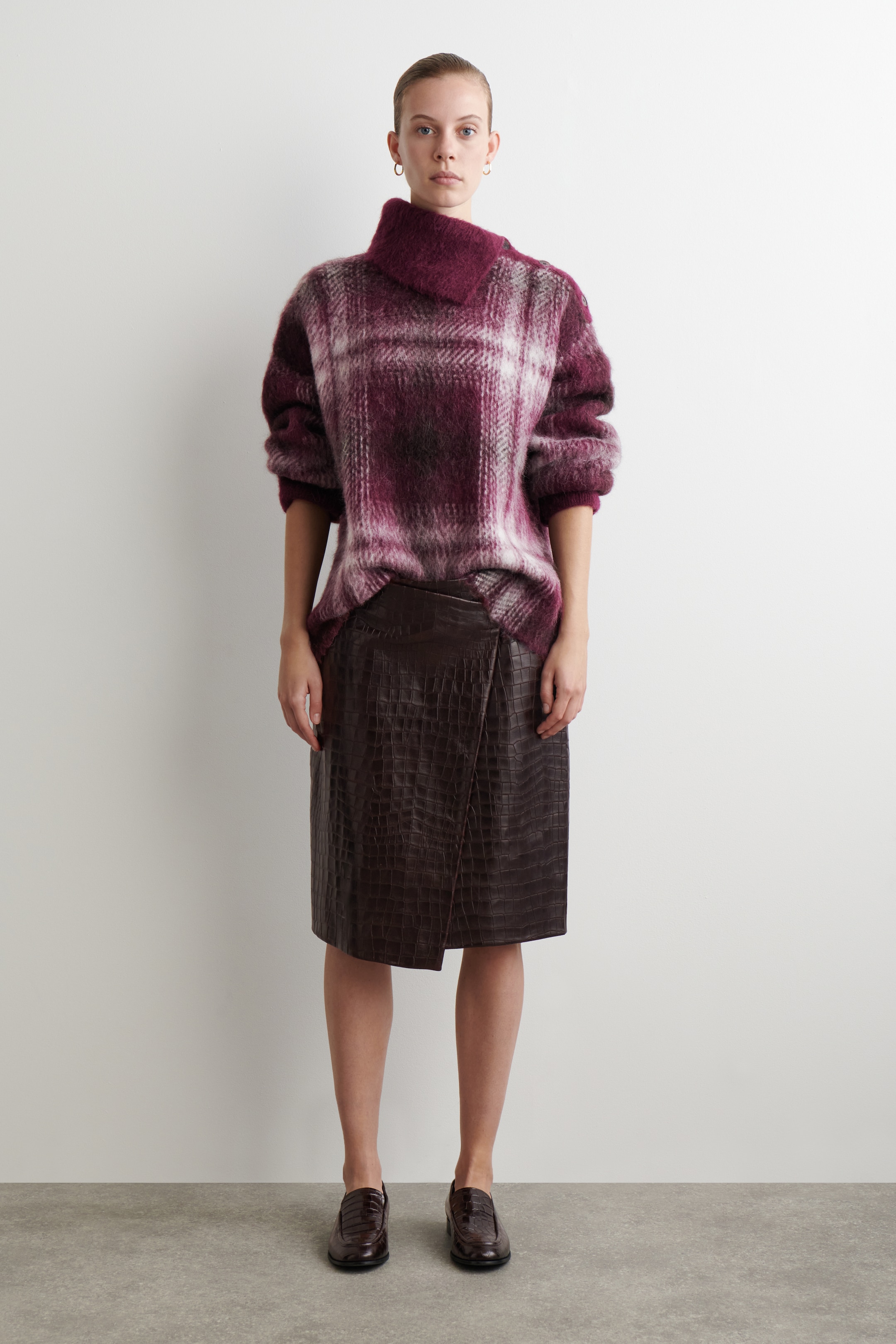 Agrandir l'image: Un pull surdimensionné et duveteux avec un motif à carreaux bordeaux, blanc et gris foncé et un col cheminée asymétrique est porté. Il est assorti d'une jupe marron foncé, longueur genou, avec une texture embossée crocodile et un devant asymétrique, ainsi que de mocassins marron foncé embossés crocodile.
