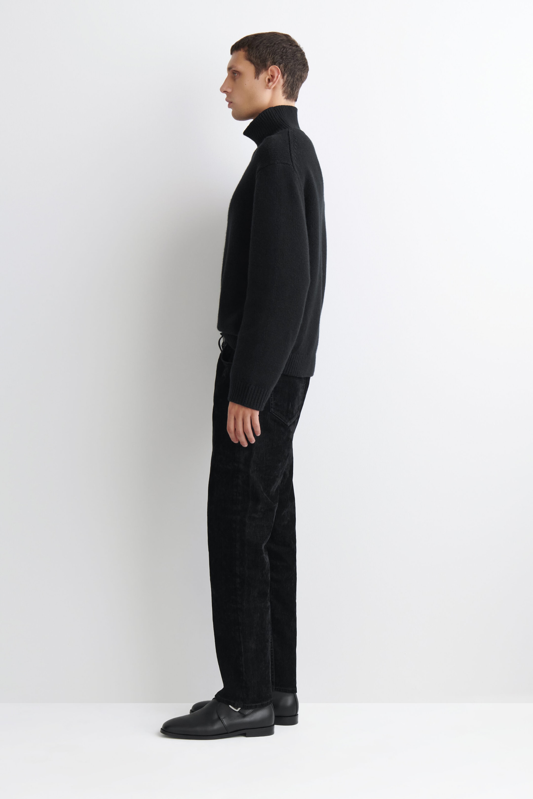 Los Signature Flocked Straight-Leg Jeans se presentan en perfil lateral orientado hacia la izquierda, destacando su silueta estructurada de pierna recta y su acabado negro flocado con textura. Los jeans caen limpiamente hasta los tobillos y se combinan con un suéter negro de cuello alto y zapatos de piel negros.