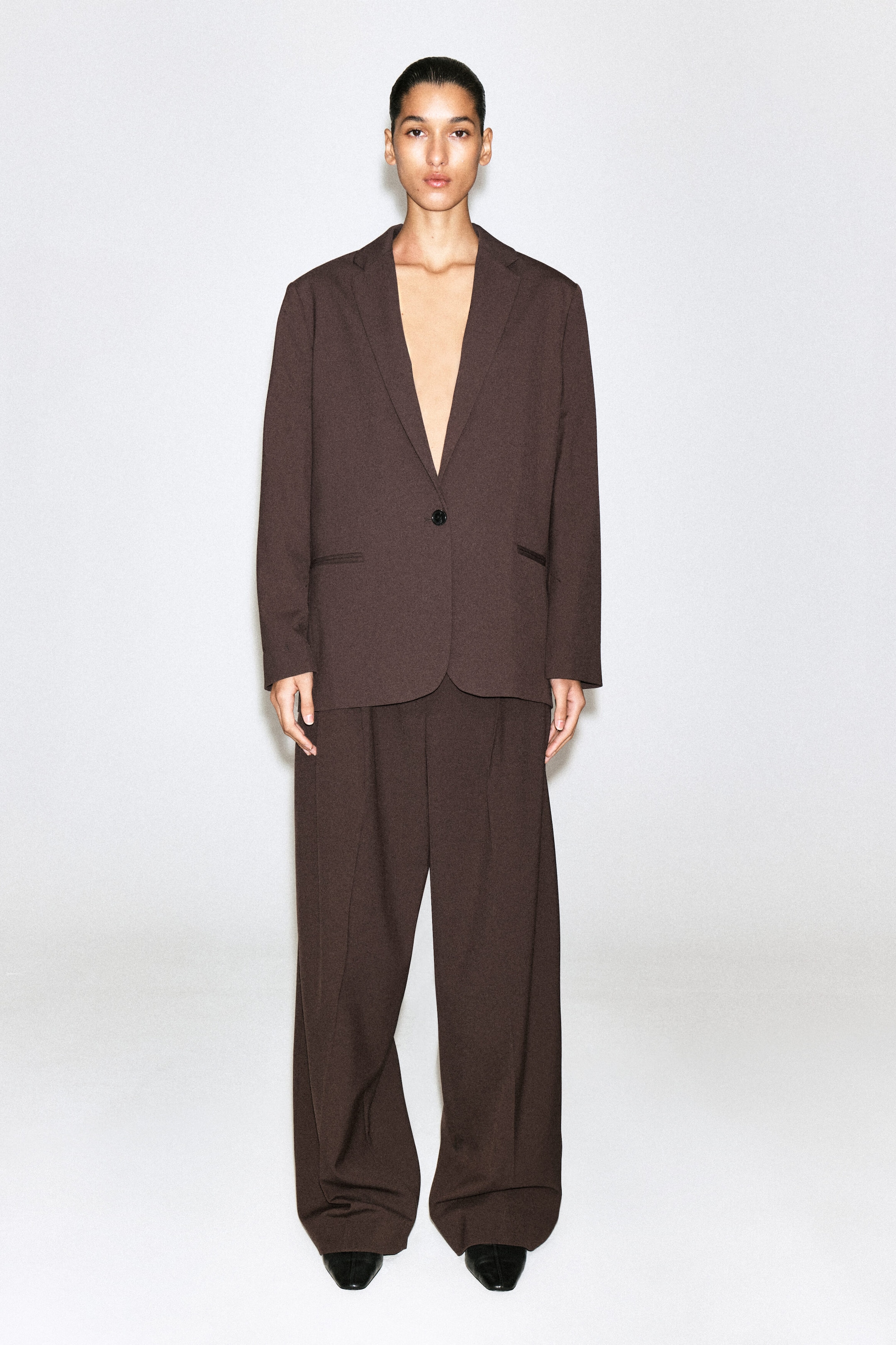 A dark brown suit is shown from the front, featuring a relaxed-fit, single-breasted blazer with notch lapels and a single-button fastening. The matching wide-leg trousers fall to pool slightly over black pointed-toe shoes.