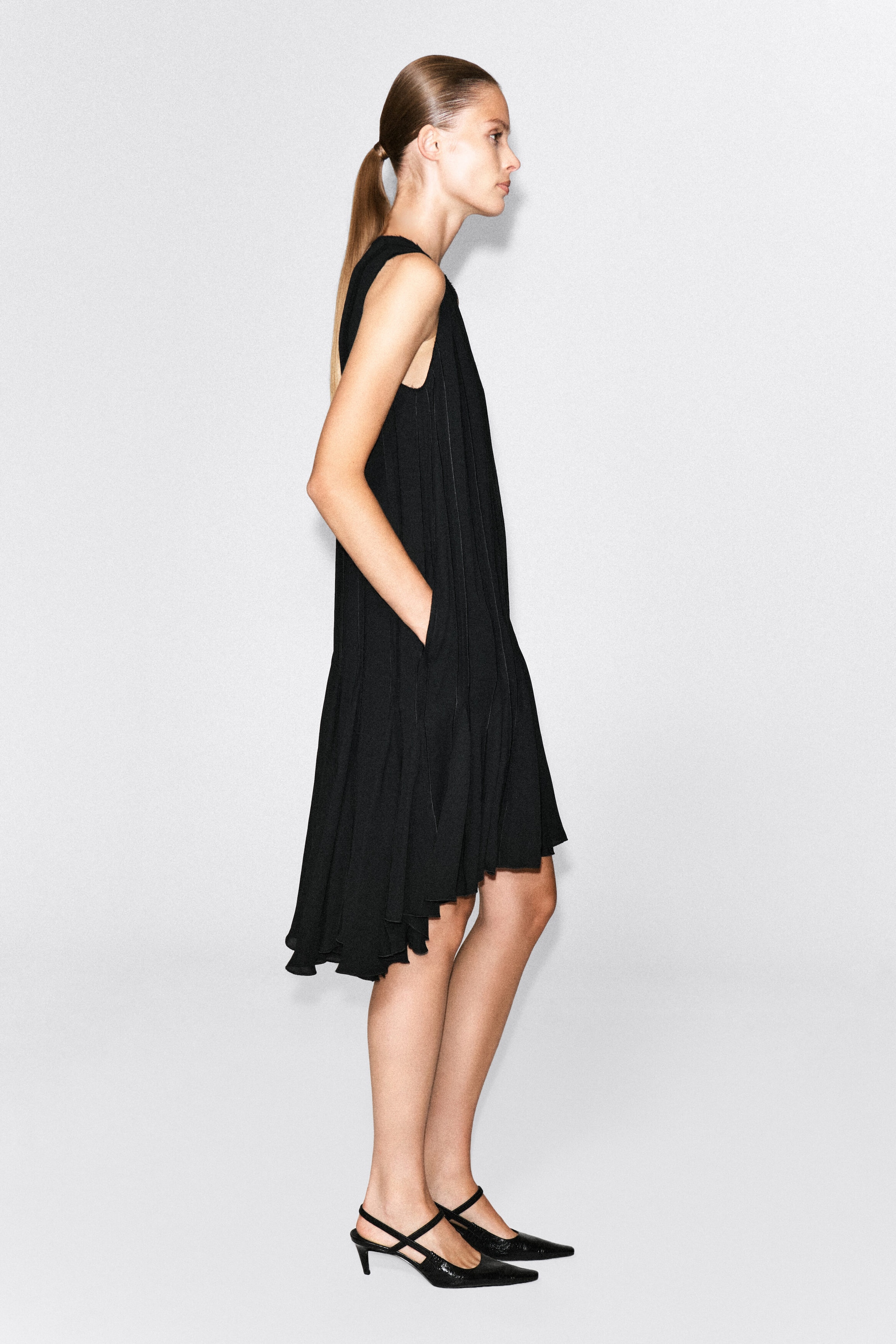 A black, sleeveless, voluminous pleated midi dress is worn by the model, who is standing in profile with one hand resting in a side pocket. The dress features an asymmetrical high-low hemline and is paired with black pointed-toe slingback heels. A black, sleeveless, voluminous pleated midi dress is worn by the model, who is standing in profile with one hand resting in a side pocket. The dress features an asymmetrical high-low hemline and is paired with black pointed-toe slingback heels.