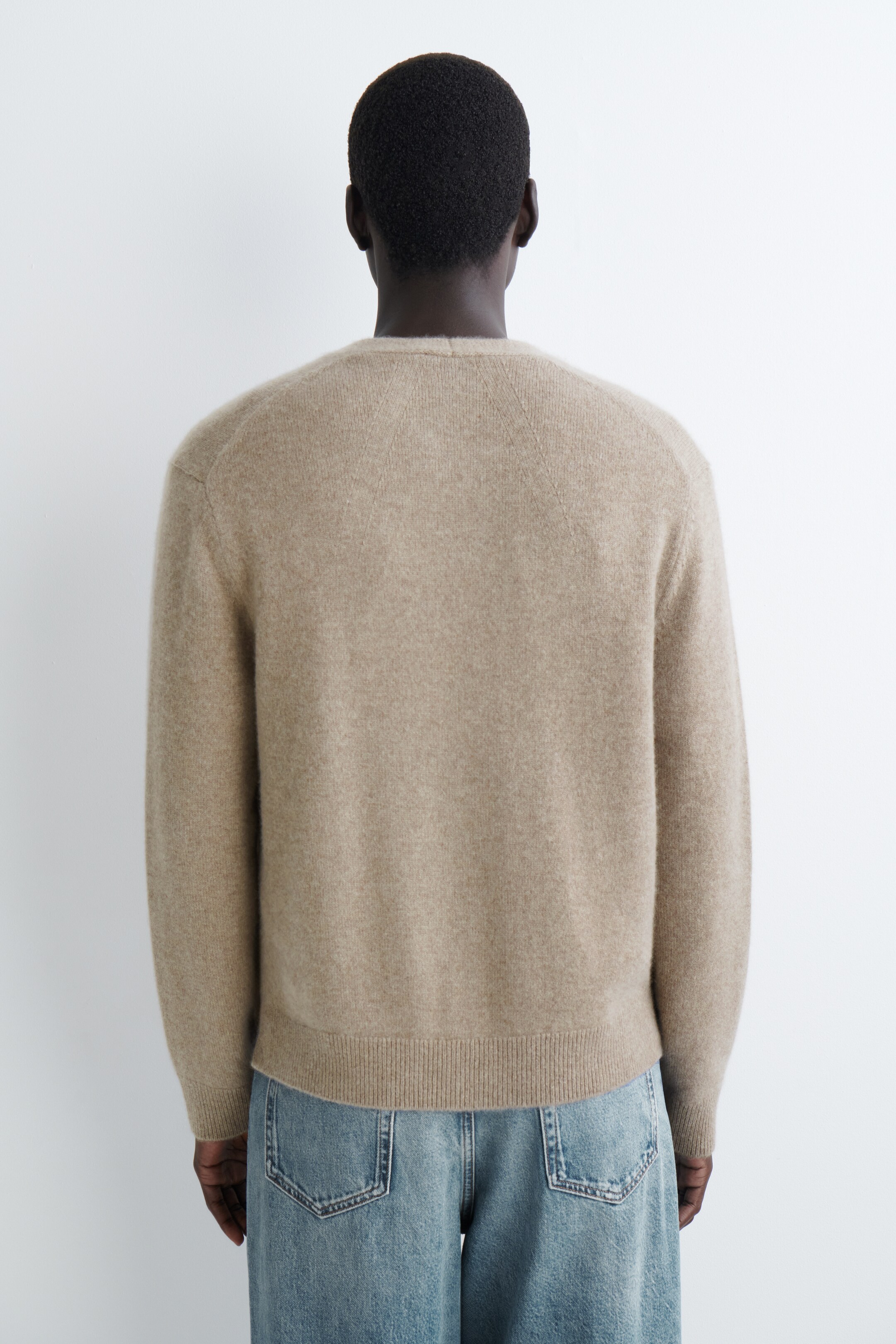 Grotere afbeelding bekijken: Een lichtbeige trui met ronde hals wordt van de achterkant gepresenteerd. Het heeft raglanmouwen en een geribde onderzoom, en wordt gedragen over lichtblauwe jeans.