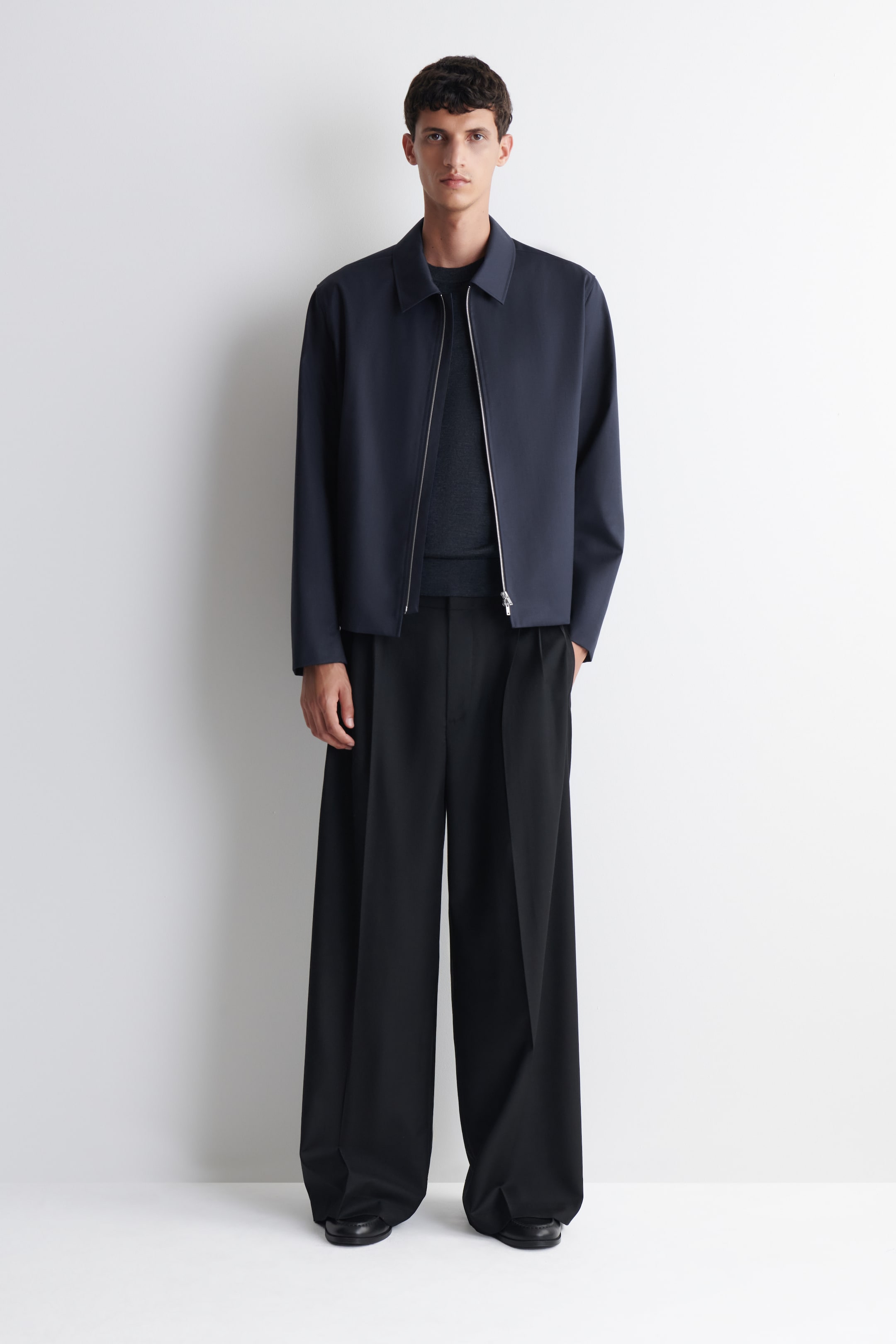 Agrandir l'image: Une veste bomber bleu marine foncé avec un col pointu et une fermeture éclair argentée est portée par le mannequin, ouverte sur un pull à col rond gris foncé. Le mannequin porte également un pantalon plissé noir à jambe large et des mocassins noirs.