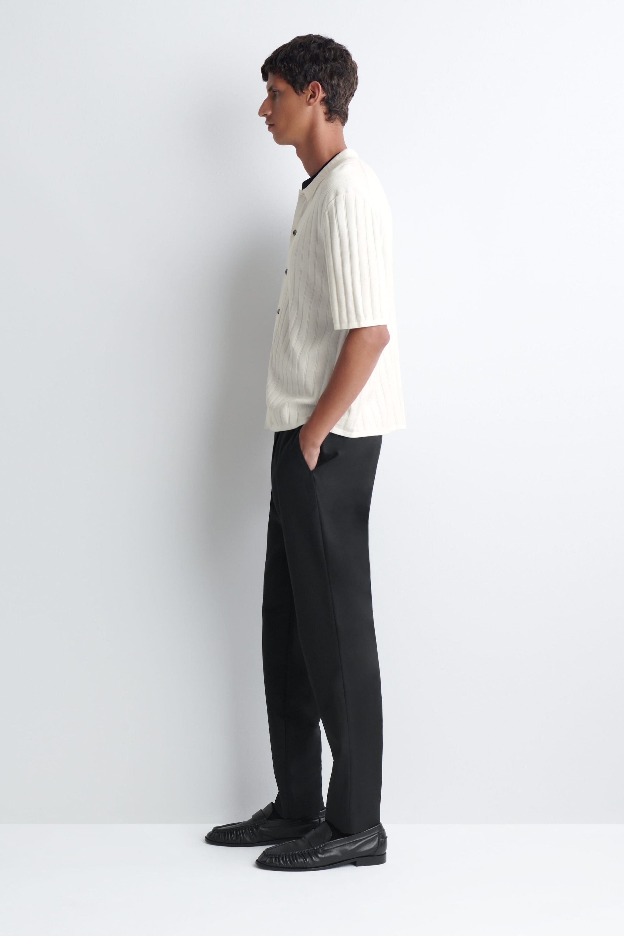 Ein weißes Baumwollhemd aus Rippstrick mit kurzen Ärmeln, Kragen und markanter vertikaler Rippstruktur wird vom Model getragen. Das Hemd mit Knopfleiste wird über einem schwarzen Kragen gestylt und mit einer schwarzen Hose sowie schwarzen Loafern kombiniert. Ein weißes Baumwollhemd aus Rippstrick mit kurzen Ärmeln, Kragen und markanter vertikaler Rippstruktur wird vom Model getragen. Das Hemd mit Knopfleiste wird über einem schwarzen Kragen gestylt und mit einer schwarzen Hose sowie schwarzen Loafern kombiniert.