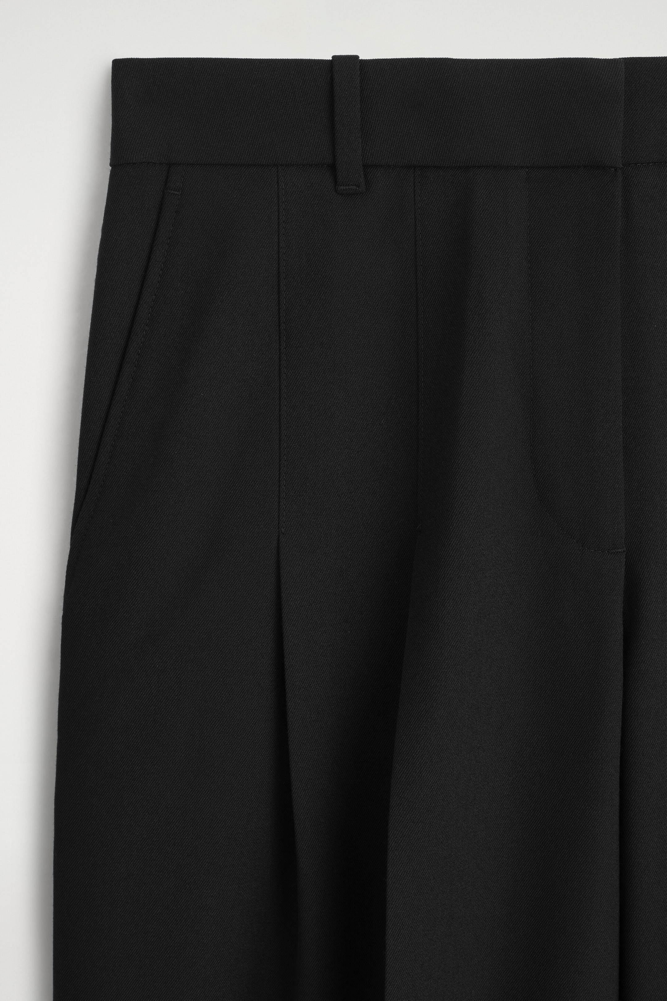 A close-up shot shows the front of the black pleated wool-blend wide-leg trousers, highlighting the structured waistband with a belt loop, a side slant pocket, and sharp front pleats draping downwards. A close-up shot shows the front of the black pleated wool-blend wide-leg trousers, highlighting the structured waistband with a belt loop, a side slant pocket, and sharp front pleats draping downwards.